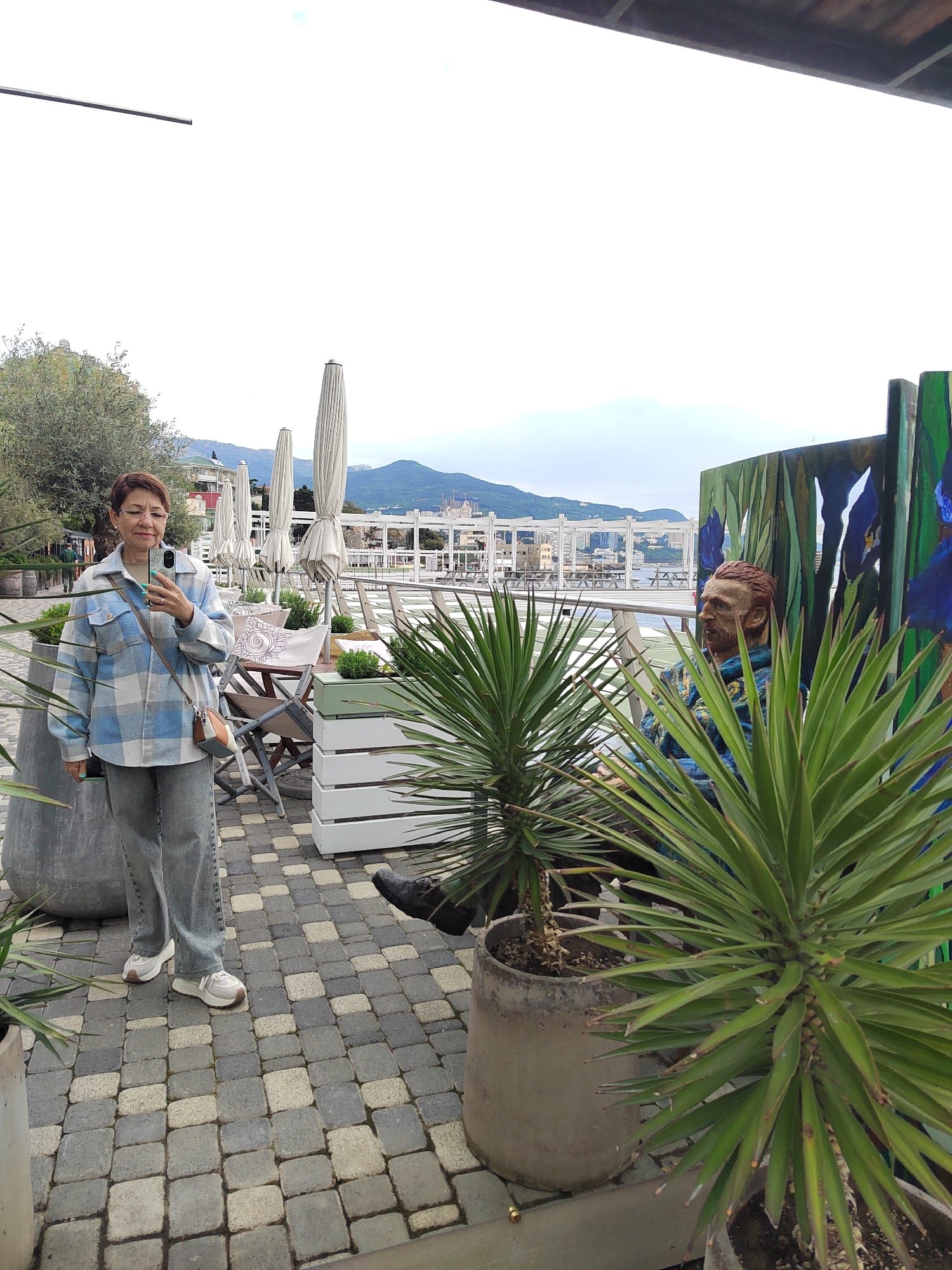 The image captures a scene in Unknown, Unknown. A middle-aged woman stands on a paved area, holding a phone as if taking a picture. She is wearing a light blue and white plaid jacket, gray jeans, and white sneakers. A crossbody bag is slung over her shoulder.
Behind her, tables and chairs with closed parasols indicate an outdoor cafe or patio setting. In the background, a body of water, a white pergola-like structure, and distant mountains are visible.
To the right, a statue resembling Vincent van Gogh sits amidst potted plants. The statue is dressed in blue clothing and placed in front of a screen painted with blue and green strokes, possibly depicting irises or foliage. The potted plants are a prominent foreground element. The overall environment suggests a tourist-friendly location with artistic elements integrated into the landscape.