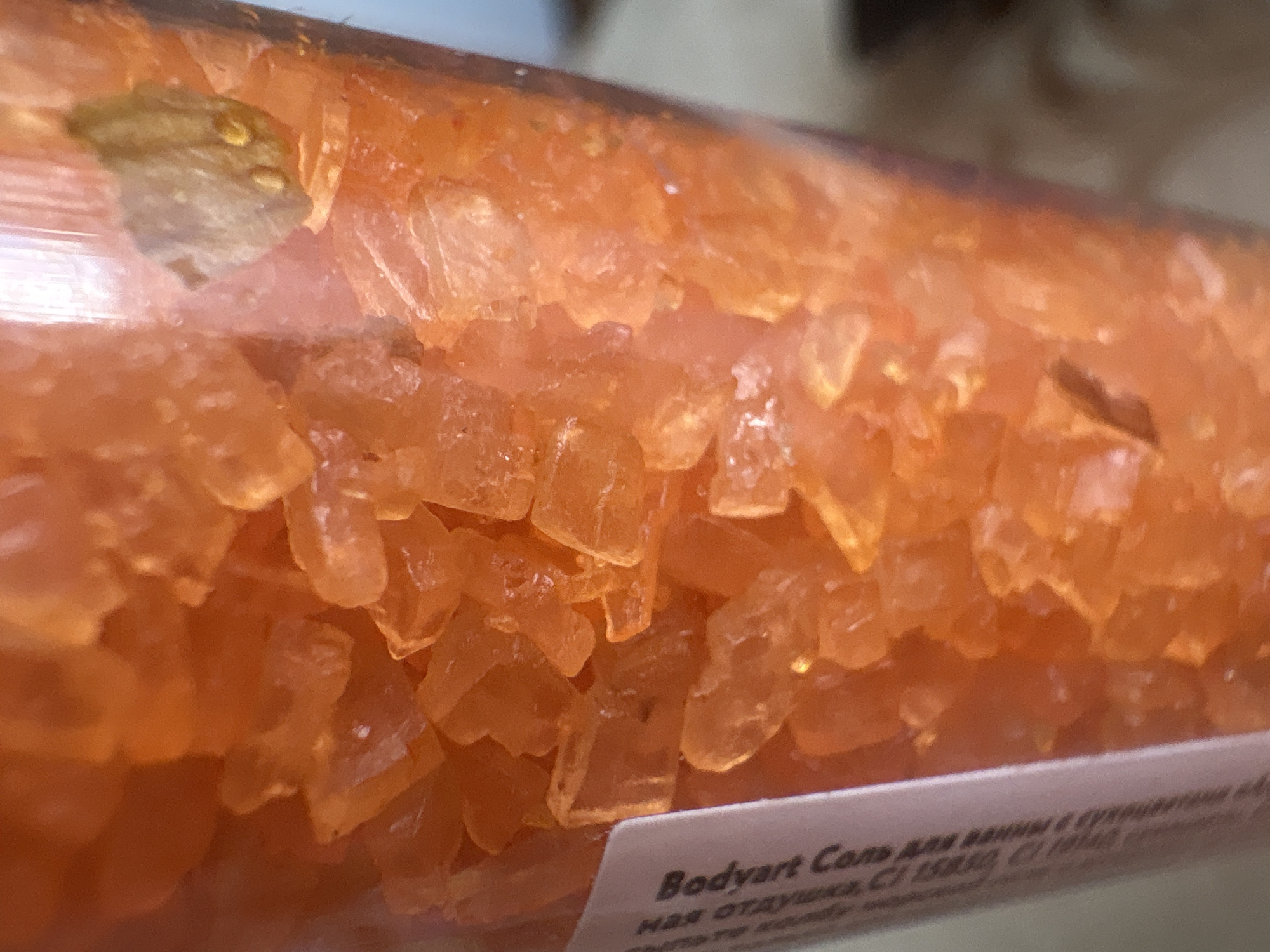 The image shows a close-up of orange bath salts inside a clear container. There is a visible label with the text "Bodyart Соль для ванны с" on it. The top of the container is visible, and there is what appears to be a dried plant element mixed in with the salts at the top left. Based on the Cyrillic text on the label (which reads "Bodyart Sol' dlya vanny s...", meaning "Bodyart Bath Salts with..."), it can be inferred the product is likely made or sold in a region where Russian is spoken. No specific location within the city of Chaikovsky, Russia, can be determined from the image.