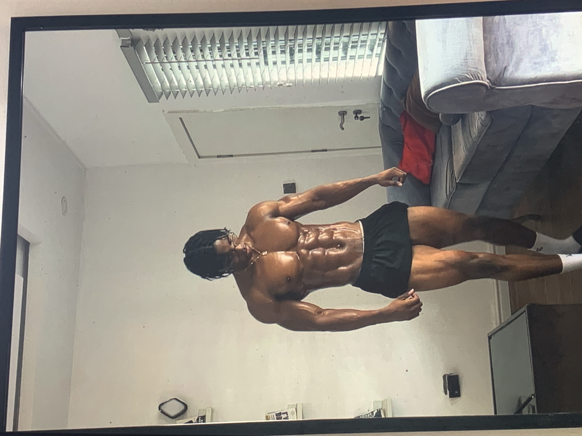 A full-body shot captured through what appears to be a mirror, shows a muscular, dark-skinned man posing indoors. The man has braided hair, is wearing glasses, a gold chain, black shorts, and white socks. His torso is bare, revealing a well-defined physique, and his skin appears shiny, possibly from oil or sweat, enhancing muscle definition in his chest, shoulders, arms, and abdominal area. He stands with his arms slightly raised and bent, showcasing his upper body.
The background reveals a simple indoor setting. To the man's right, a large gray sofa or couch with decorative piping is visible. A red pillow is partially seen tucked behind the sofa cushions. Above and behind the man, a ceiling with a rectangular fluorescent light fixture, featuring a slatted diffuser, is prominent. Further back, a light-colored door or opening with a door handle or hook is visible. The walls are plain and light-colored. Towards the bottom right, a dark, rectangular object, possibly a monitor or piece of furniture, is present. Several small, indistinct items are scattered on a surface near the bottom edge of the frame. The overall impression is a personal fitness display within a residential or private indoor space. No discernible text is visible.
