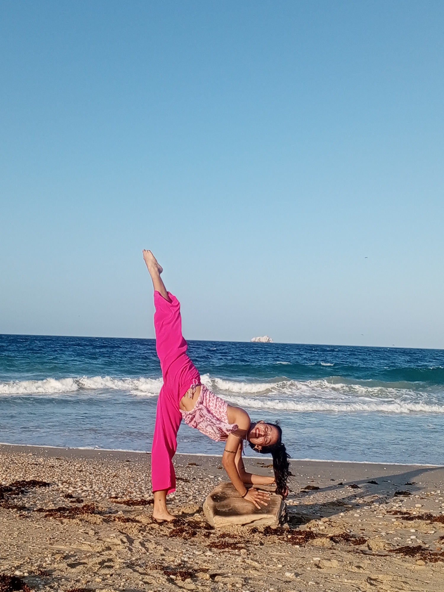 A woman is performing a yoga pose on a beach in Porlamar, Venezuela. She is wearing pink pants and a patterned top. Her left leg is extended upwards, and her hands are placed on a rock. The ocean is behind her, with small waves reaching the shore. The sky is clear and blue. The beach sand is light brown and has pieces of seaweed scattered around the woman. In the distance, there is a small island.