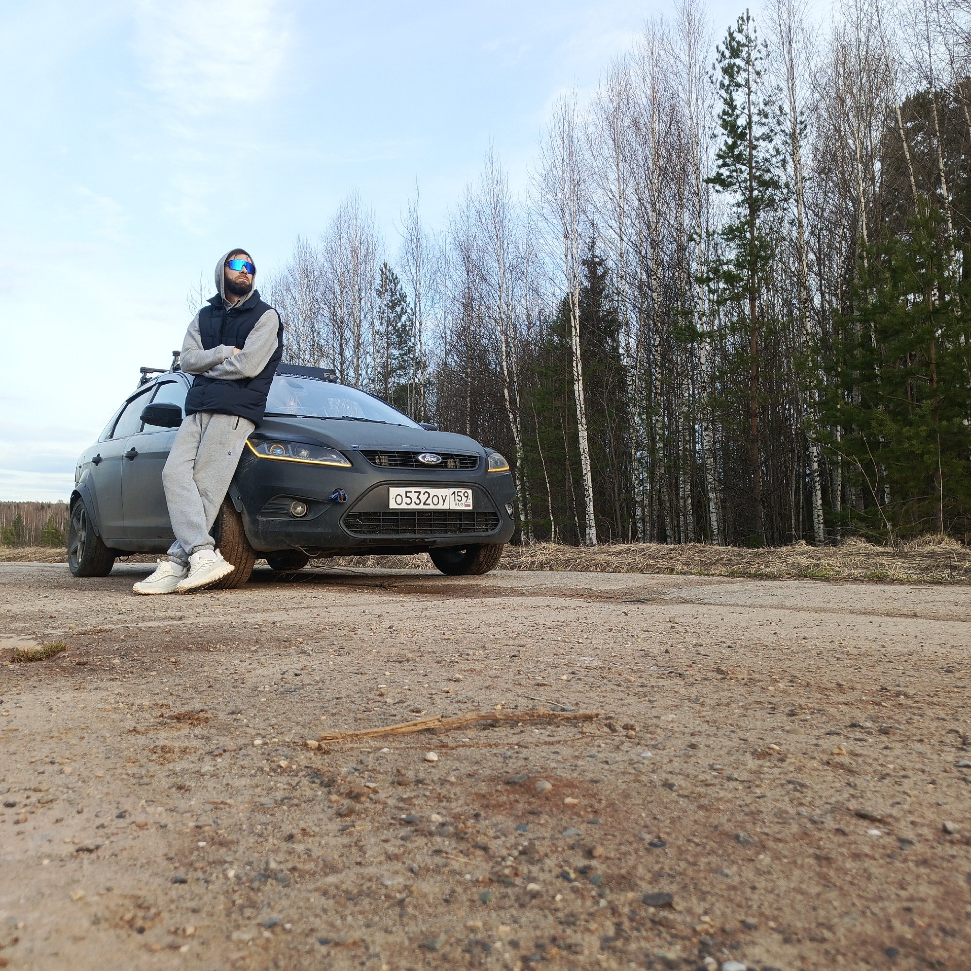 A man wearing a gray hooded sweatshirt, black vest, gray sweatpants, white sneakers, and blue sunglasses stands leaning against the front of a black Ford Focus. The car is parked on a dirt road, and its license plate reads "0532OY 159." In the background, a forest of mostly birch trees is visible under a partly cloudy sky. The location is Марковский, Russia.