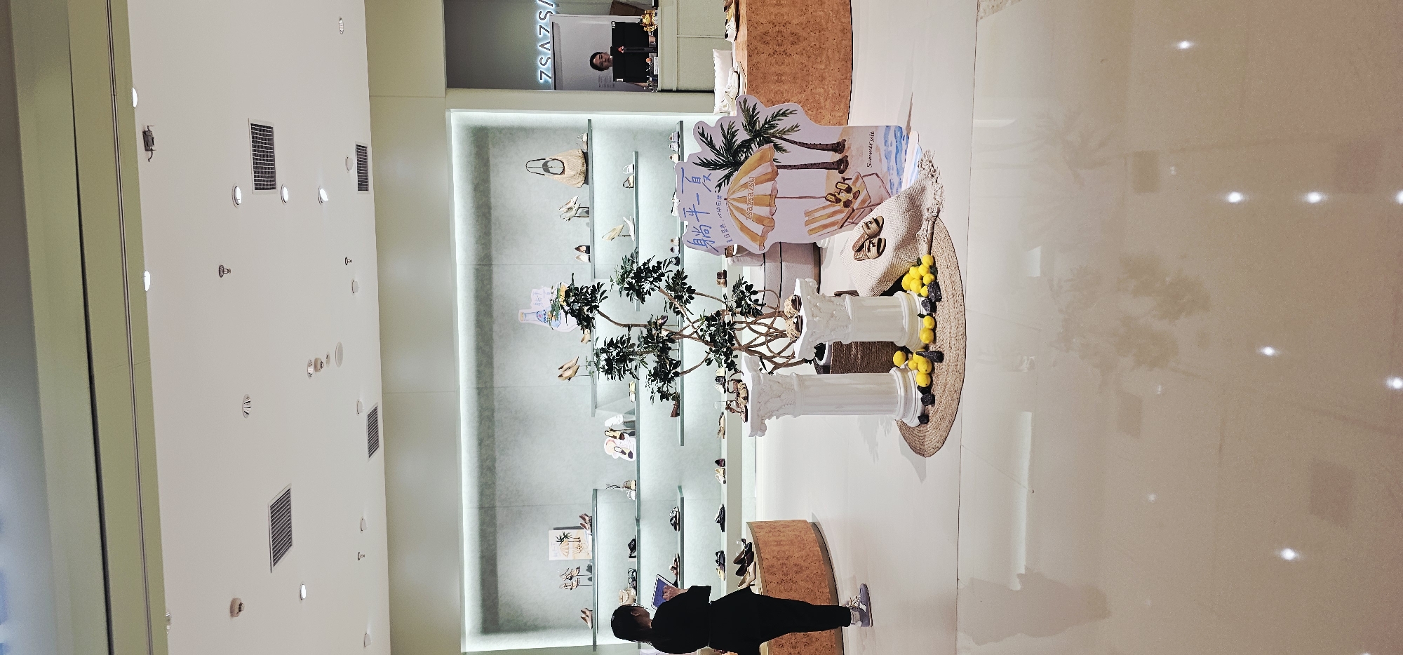 This is a photograph taken at a shoe store in Jiulongpo District, China. The store features light green walls and shelves displaying various shoes and handbags. A woman in a black shirt and black pants is standing on the left side of the image, looking at a tablet. In the center, a decorative display includes two white pillars, a potted plant, a woven mat, lemons, and stones. To the right of the display is a cardboard cutout advertising a summer sale, depicting a beach scene with an umbrella and palm trees. A person is visible behind a counter on the far right of the image, possibly a cashier. The floor is a light-colored, reflective surface.