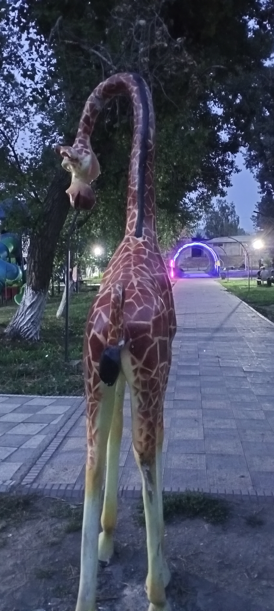 A statue of a giraffe is the primary subject in the foreground of this outdoor image, which appears to be taken at dusk in the city of Sokuluk, Kyrgyzstan. The giraffe statue is viewed from behind, with its long neck curved downwards, revealing its head and face. The statue is detailed with the characteristic brown and beige patterned coat of a giraffe.
The giraffe statue stands on a paved pathway made of grey rectangular stones, bordered by grass. To the left of the pathway, there is a grassy area with a large tree and playground equipment, including blue and green slides. Further down the pathway, a brightly lit, archway-like structure with blue and pink neon lights is visible in the distance.
Several streetlights illuminate the pathway and surrounding areas. In the background, there are some buildings and trees, and a few people are seated on benches on the right side of the pathway. The sky is a dusky blue, suggesting the late evening or early morning.