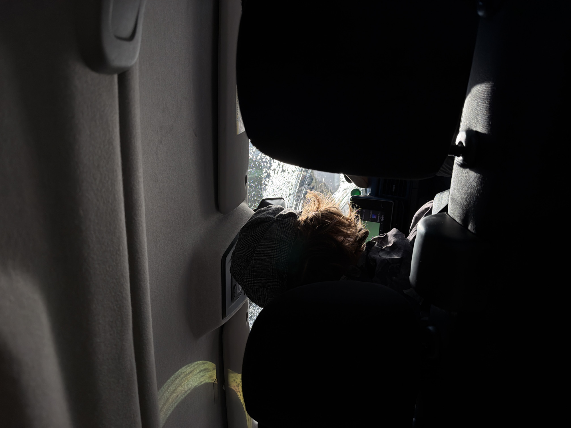 A person with reddish-brown hair and a gray hat appears to be sleeping in the back of a vehicle, with their head resting against the window. The window has a speckled appearance, possibly due to water droplets. The vehicle interior consists of a light gray wall and dark seats. The lighting is uneven, with bright sunlight filtering through the window and casting shadows. No specific details indicate the location is Saint-André, France.
