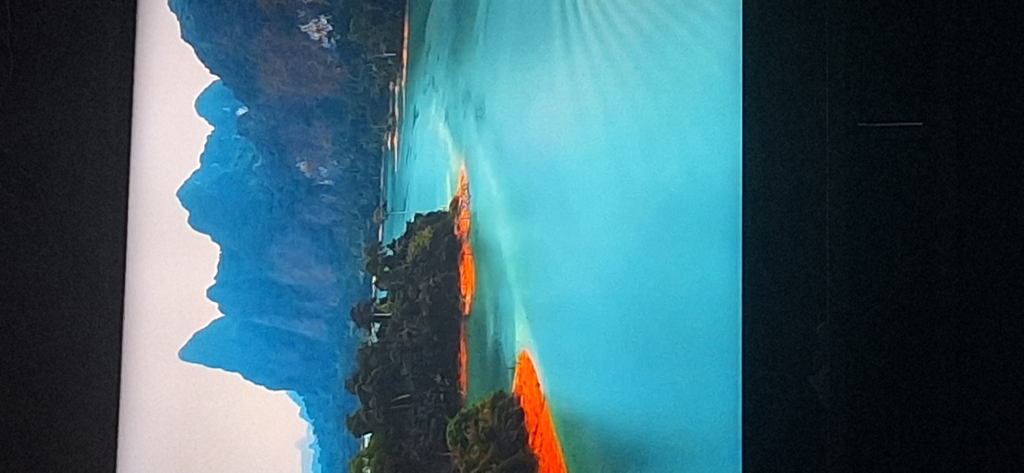 This image depicts a striking natural landscape, not a scene within the city of Bila Tserkva, Ukraine. The photograph captures a body of turquoise water, likely a lake or a wide river, with distinctive reddish-orange shoreline areas. Lush, dark green vegetation covers a peninsular landmass jutting into the water. In the background, a range of jagged, blue-hued mountains rises against a pale sky. The overall impression is one of a remote and scenic natural environment, possibly a tropical or subtropical location, with no discernible elements connecting it to the specified Ukrainian city.