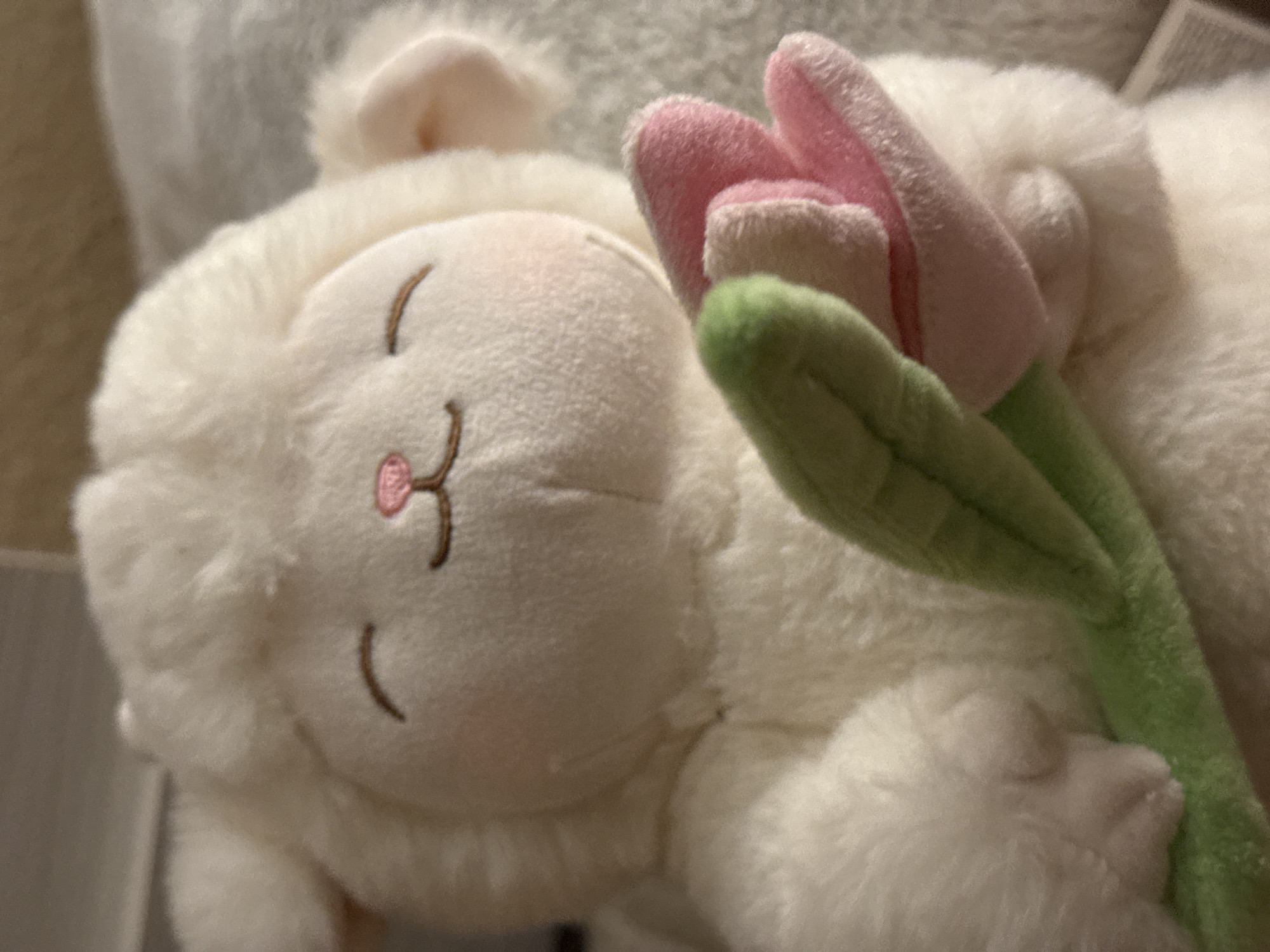 The image shows a close-up of a fluffy white lamb stuffed animal holding a bouquet of three plush tulips. The lamb has closed eyes with subtle brown stitching, a small pink nose, and rosy cheeks. The tulips are a mix of pink and green. The stuffed animal is resting on a soft, textured surface, possibly a blanket or rug, with a neutral background. There is no discernible location in Tbilisi, Georgia.