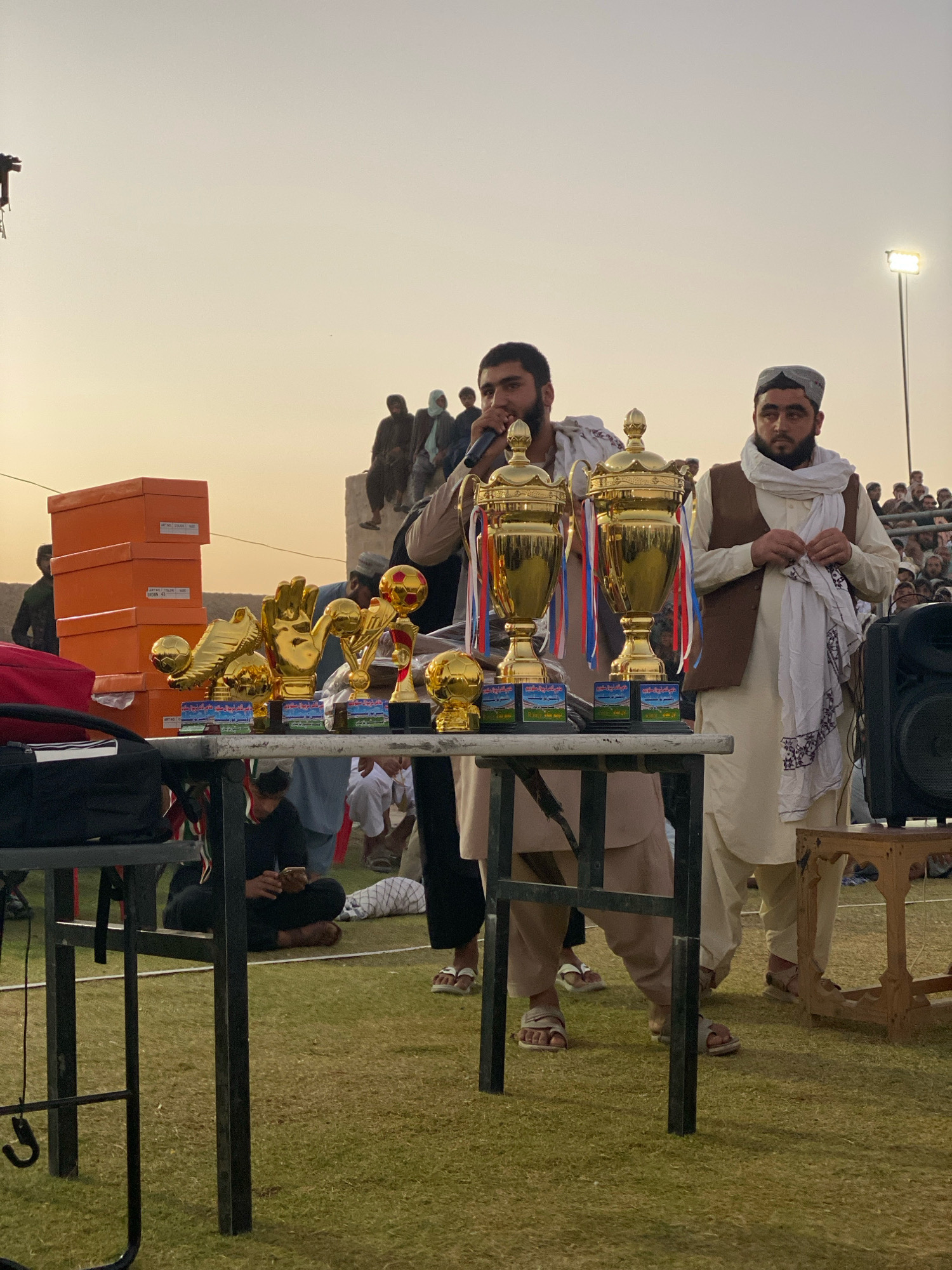 The image depicts an outdoor event in Spin Boldak, Afghanistan, with a group of men standing around a table displaying trophies and orange boxes. Two men in the foreground are holding microphones and appear to be addressing the crowd. One man, wearing a light-colored shalwar kameez and a brown vest, is holding a microphone and speaking. Next to him, another man, also in traditional clothing and a white scarf, is looking towards the trophies.
The table is laden with several gold-colored trophies, some shaped like soccer balls and a large glove. Behind the table, a stack of orange boxes is visible, along with other individuals in traditional attire. The background shows a crowd of people seated and standing, with a lamppost in the distance. The event appears to be taking place on a grassy field, possibly a sports ground. The sky is overcast with a warm, diffused light, suggesting it could be early morning or late afternoon.