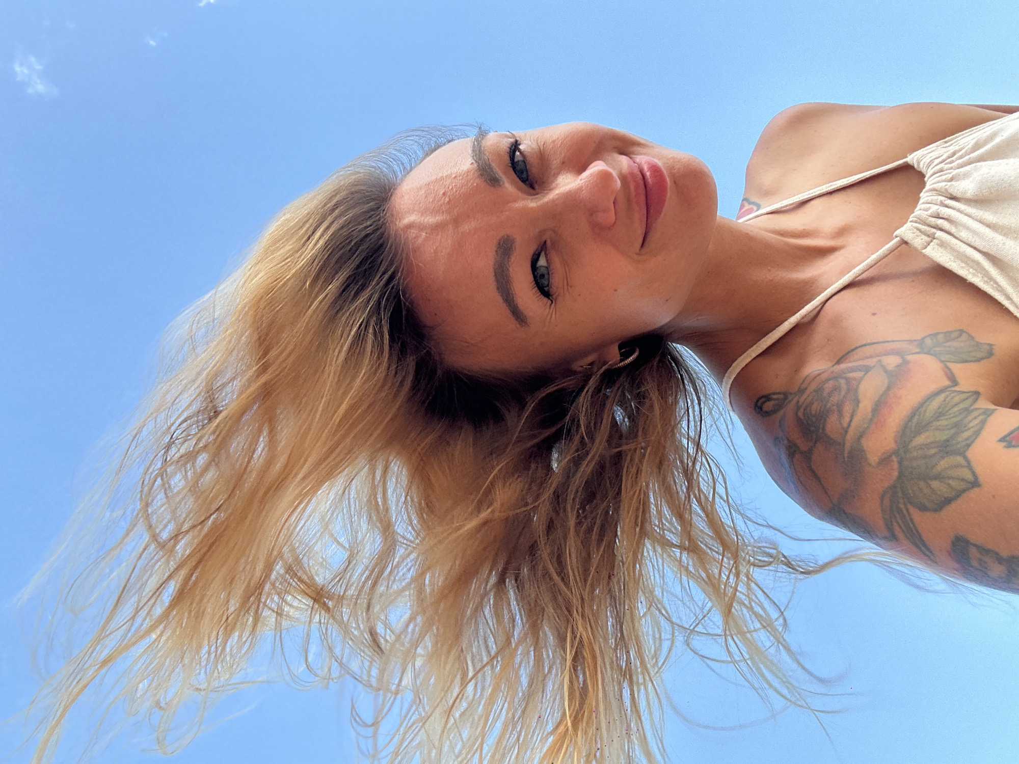 A woman is captured from a low-angle perspective, looking directly down at the camera against a clear, bright blue sky. Her head is tilted slightly to her right, and she displays a soft, engaging smile. She has long, blonde to light brown hair that appears wind-swept, fanning out around her head. Her blue eyes are open and focused, and her eyebrows are distinctly shaped.
She is wearing a light-colored, possibly beige or off-white, top with thin straps and gathered fabric at the bust. Several tattoos are visible: a large floral design featuring roses and leaves covers her right shoulder and upper arm, and a small red tattoo is visible on her upper chest/collarbone area. A hoop earring is present in her right ear. The background is a vibrant, cloudless blue sky, indicating a sunny outdoor setting during the daytime. The overall impression is one of a relaxed and joyful moment. The location context for this image is Mũi Né, Vietnam. There is no visible text in the image.