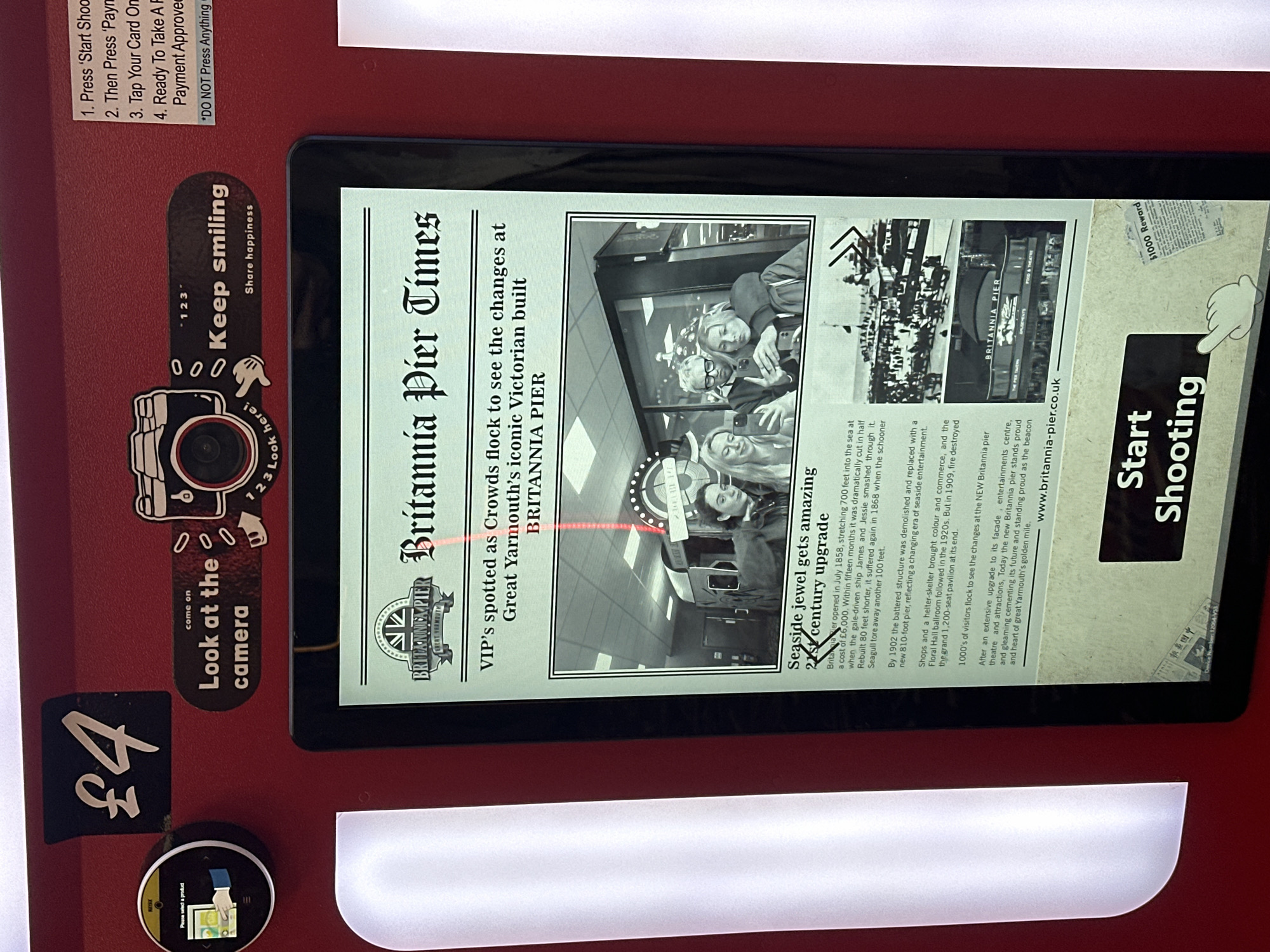 A red digital photo booth, likely situated indoors, is presented with an interactive display. The upper left section displays "£4" in black text on a white cutout. Adjacent to this is a circular black digital interface showing "Please select a product" with a graphic of a hand interacting with a screen. Centrally positioned above the main screen is a white sticker with a black camera icon and instructions: "Look at the camera" and "Keep smiling," with "Share happiness" below. Further to the right, a white label lists operating instructions: "1. Press 'Start Shooting'", "2. Then Press 'Payment'", "3. Tap Your Card Once", "4. Ready To Take A Photo! Payment Approved!", followed by a partial warning "*DO NOT Press Anything".
The large central screen displays a digital newspaper-style interface titled "Britannia Pier Times," featuring the "Britannia Pier Great Yarmouth" logo. A headline reads: "VIP's spotted as Crowds flock to see the changes at Great Yarmouth's iconic Victorian built BRITANNIA PIER". Below this, a black and white image shows three young individuals—two females and one male wearing glasses—posing for a selfie with a smartphone, likely a reflection of people standing in front of the booth. The male is in the center with a female on either side, one holding the phone. Beneath this image, an article titled "Seaside jewel gets amazing 21st century upgrade" details the history of Britannia Pier in Great Yarmouth, mentioning its 1858 opening, reconstruction after a 1909 fire, and modern updates. The article includes smaller historical photographs of the pier and a web address, "www.britannia-pier.co.uk". At the bottom center of the screen, a prominent black button with white text reads "Start Shooting," overlaid with a faint, transparent hand icon pointing towards it. To the right of this button, a partially obscured "$1000 Reward" notice is visible. The booth's red casing and the screen reflect ambient light sources, indicating a well-lit environment.