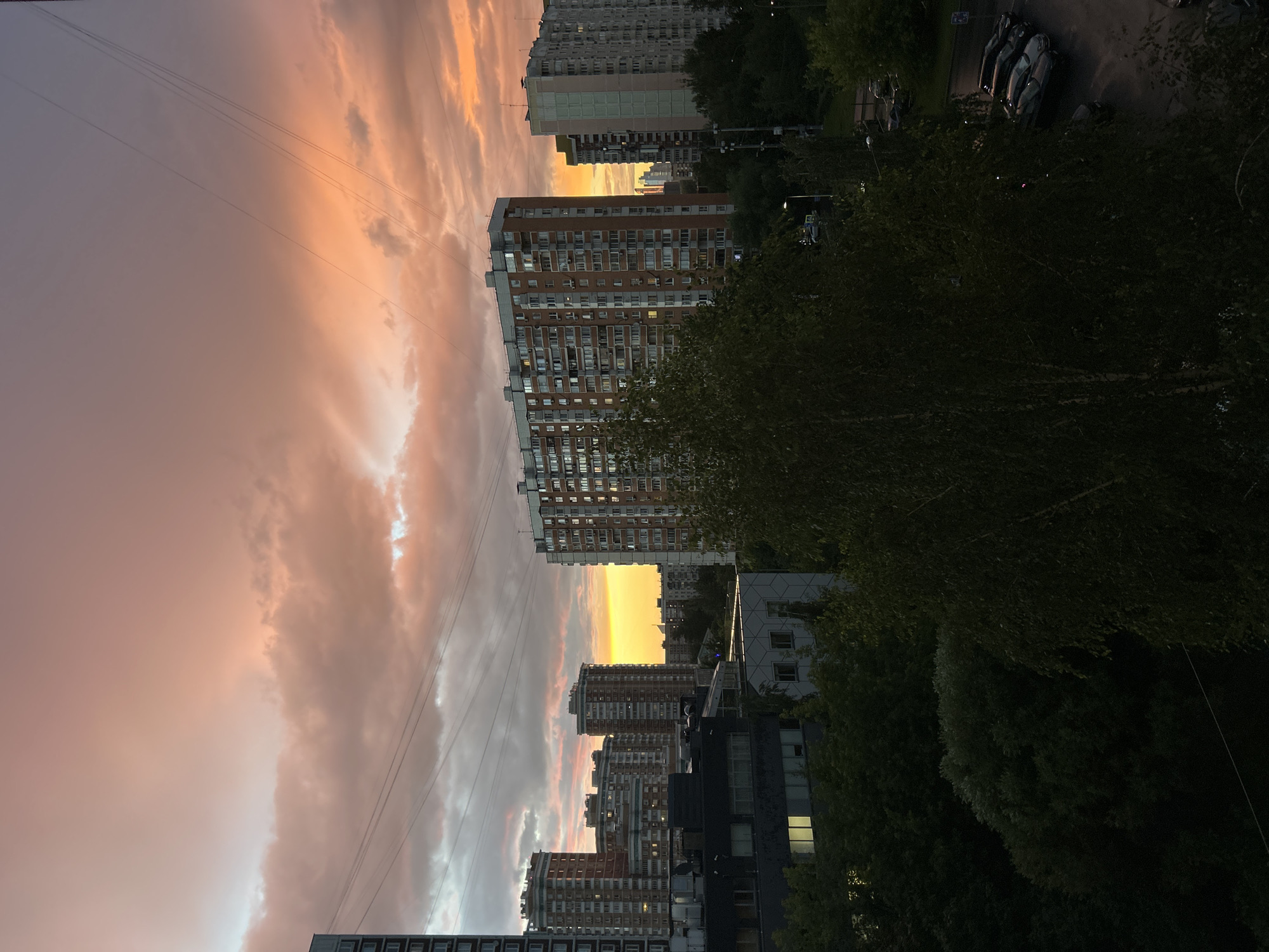 The image depicts a cityscape at sunset in Moscow, Russia. The primary subjects are several tall apartment buildings and a vibrant sky filled with clouds illuminated by the setting sun. The sky is a dominant feature, showcasing a gradient of colors from a deep purple-gray at the top left to warm oranges and pinks as it moves towards the horizon, with streaks of white light breaking through the clouds. The buildings are of varying architectural styles, mostly modern high-rise residential blocks with numerous windows.
The setting appears to be an urban residential area, likely a densely populated neighborhood. The time of day is clearly indicated by the sunset, which bathes the scene in warm, dramatic light. The weather seems to be cloudy, but not rainy, with the clouds playing a crucial role in coloring the sky.
Notable details include the presence of power lines stretching across the sky, a common sight in urban environments. In the foreground and mid-ground, lush green trees partially obscure some of the buildings, suggesting a mix of urban development and green spaces. On the right side of the image, a street is visible with parked cars, indicating a functioning urban street. One building has lights on in some of its windows, suggesting that people are home and continuing their evening routines.
The overall emotion evoked by the image is one of tranquility and beauty, with the dramatic sunset providing a sense of awe over the everyday urban landscape. There is no visible text in the image.