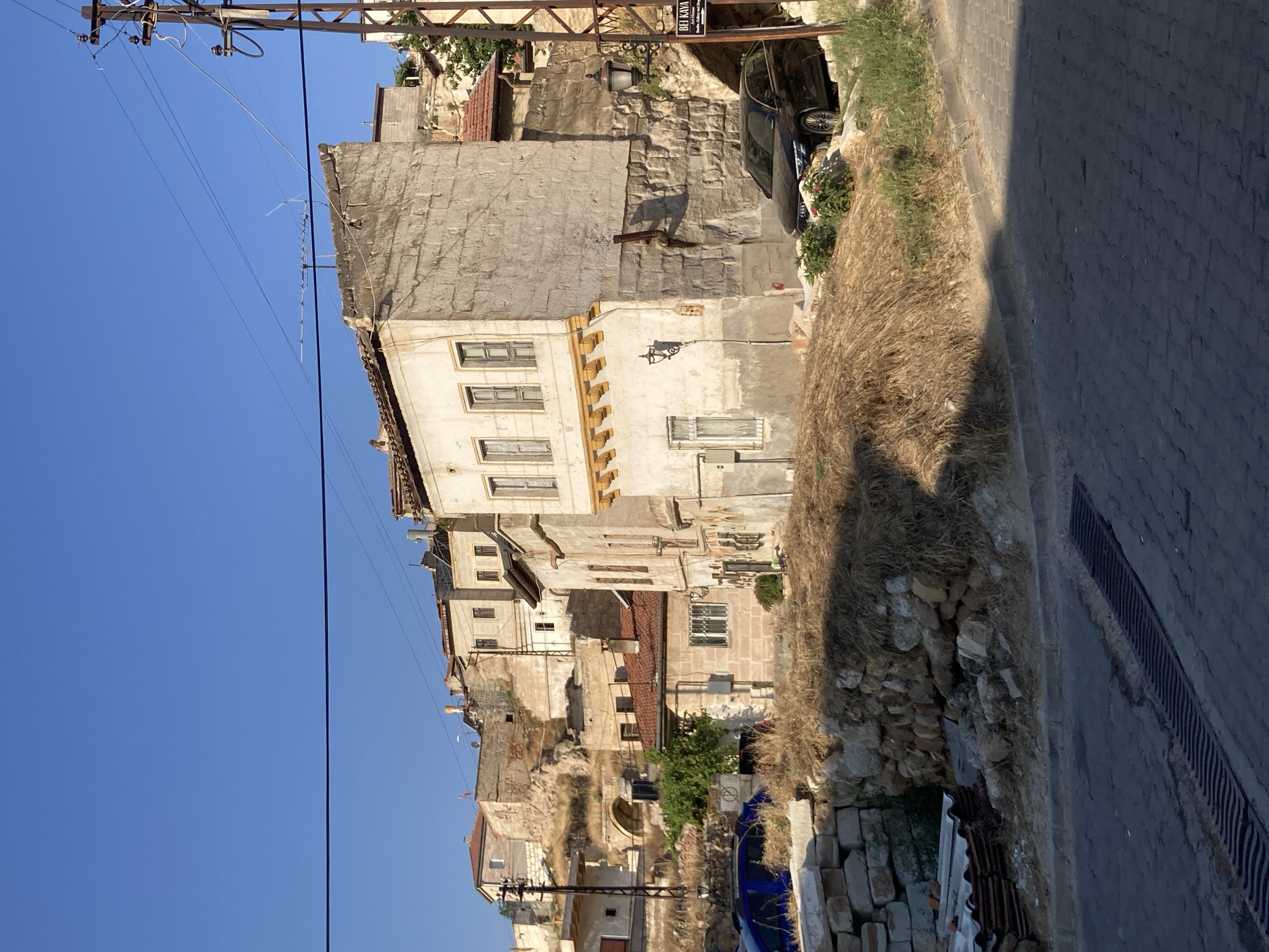 The image depicts a street scene in Avanos İlçe Merkezi, Turkey, characterized by traditional stone and stucco buildings integrated into a hillside. The architecture features multiple stories, arched doorways, and small windows, some with wooden shutters. The buildings appear weathered, with some showing signs of disrepair and exposed stonework.
In the foreground, a paved road with a drainage grate runs diagonally across the frame. To the left of the road, a pile of stacked white bricks and corrugated roofing materials sits next to a rough stone wall. Further up the slope, a blue vehicle is partially visible behind the wall.
Dominating the center of the image is a multi-story building with a light-colored stone facade. It has several windows on different levels and decorative yellow trim below the upper windows. Adjacent to this building, a rough stone wall forms part of the hillside. A black sedan is parked on the road in front of this wall.
The hillside is sparsely vegetated with dry grass and a few small trees. A wooden utility pole with electrical wires stretches across the upper part of the scene. A sign with Turkish text, possibly an advertisement for a shop or establishment, is attached to a lamp post on the right side of the frame. The sky is clear and blue, suggesting a sunny day. The overall impression is of an old, rustic urban environment.