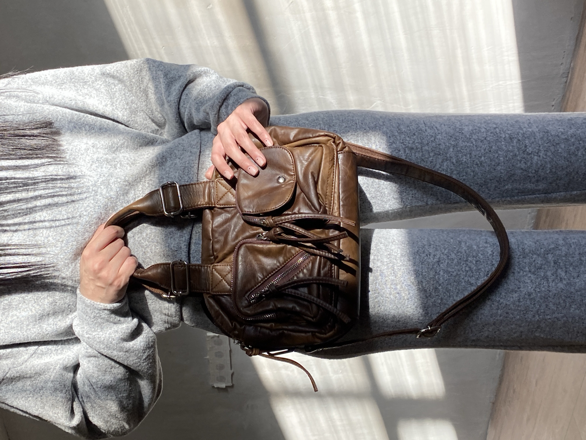 A person wearing a gray sweatsuit sits on a gray surface with a brown leather shoulder bag resting on their lap. The bag has a top flap pocket with a snap closure, a front zippered pocket, and multiple straps and buckles. The scene is bathed in sunlight. The location is unknown and there is no evidence of Votkinsk, Russia.