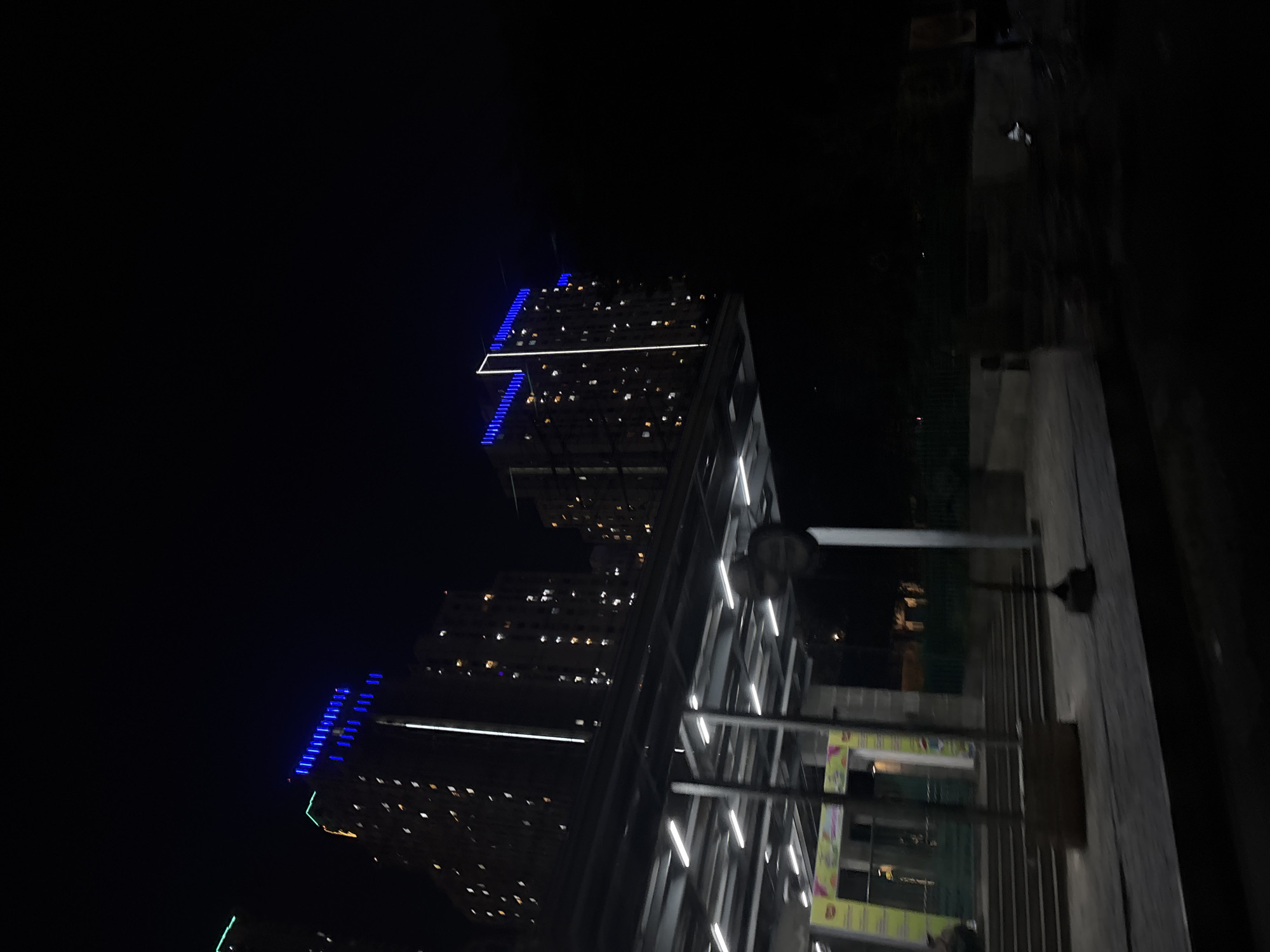 The image, taken at night in North Jakarta, Indonesia, shows a view of illuminated skyscrapers and a modern structure. The skyscrapers feature blue LED lighting along their edges and are interspersed with lit windows. In the foreground, a modern, brightly lit structure with a grid-like pattern is visible, possibly part of a building or complex. A street lamp is also visible in the foreground. The overall environment is dark, with the light sources emphasizing the buildings and structure.