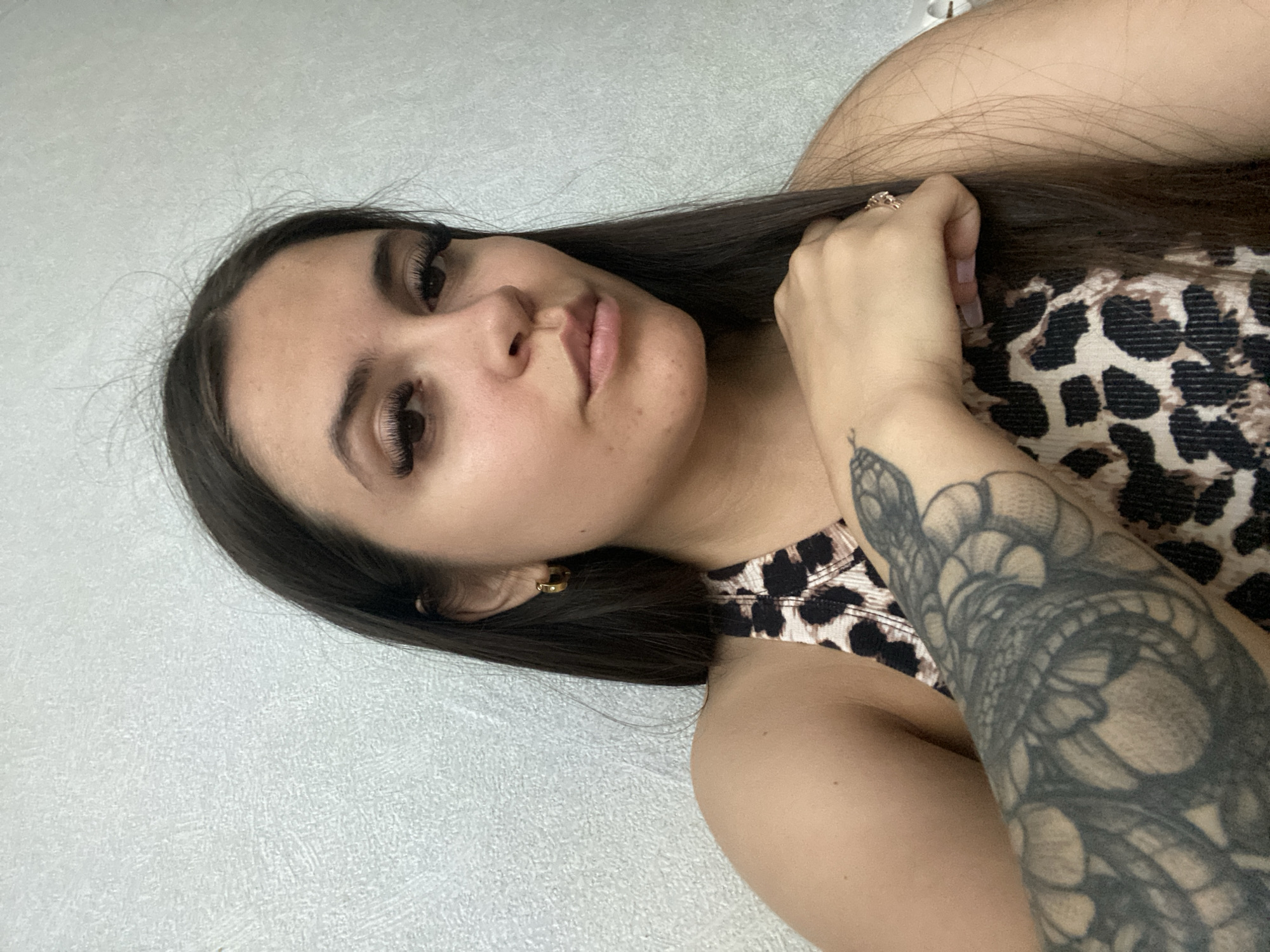 A young woman with long, dark hair and a leopard-print top is shown from the shoulders up. She is wearing hoop earrings and a ring. She has a large tattoo of a snake on her left arm. The background is a textured, light-colored wall. There is no visible indication of the location being Chaikovsky, Russia.