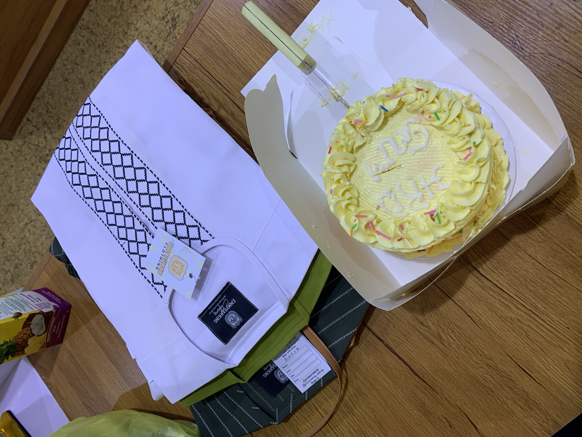 In Gura Top, Nigeria, on a wooden table, a cake and garments are displayed. A small, round cake with yellow frosting and a piped border is centrally positioned in a white cardboard box. The cake is decorated with pastel-colored sprinkles and white icing that spells out "Happy Birthday." A silver cake server with a yellow handle is partially inserted into the cake. To the left of the cake are folded garments. A white long-sleeved shirt with black geometric embroidery along the neckline and cuffs is laid out flat. A dark green striped garment is folded beneath it, with a tag visible that reads "EMMYBANKING CLOTHING." Another garment, possibly a brown bag, is partially visible underneath. In the background, a box of "Pineapple & Coconut" juice is situated on a light-colored surface. The overall scene suggests a celebratory occasion, likely a birthday.