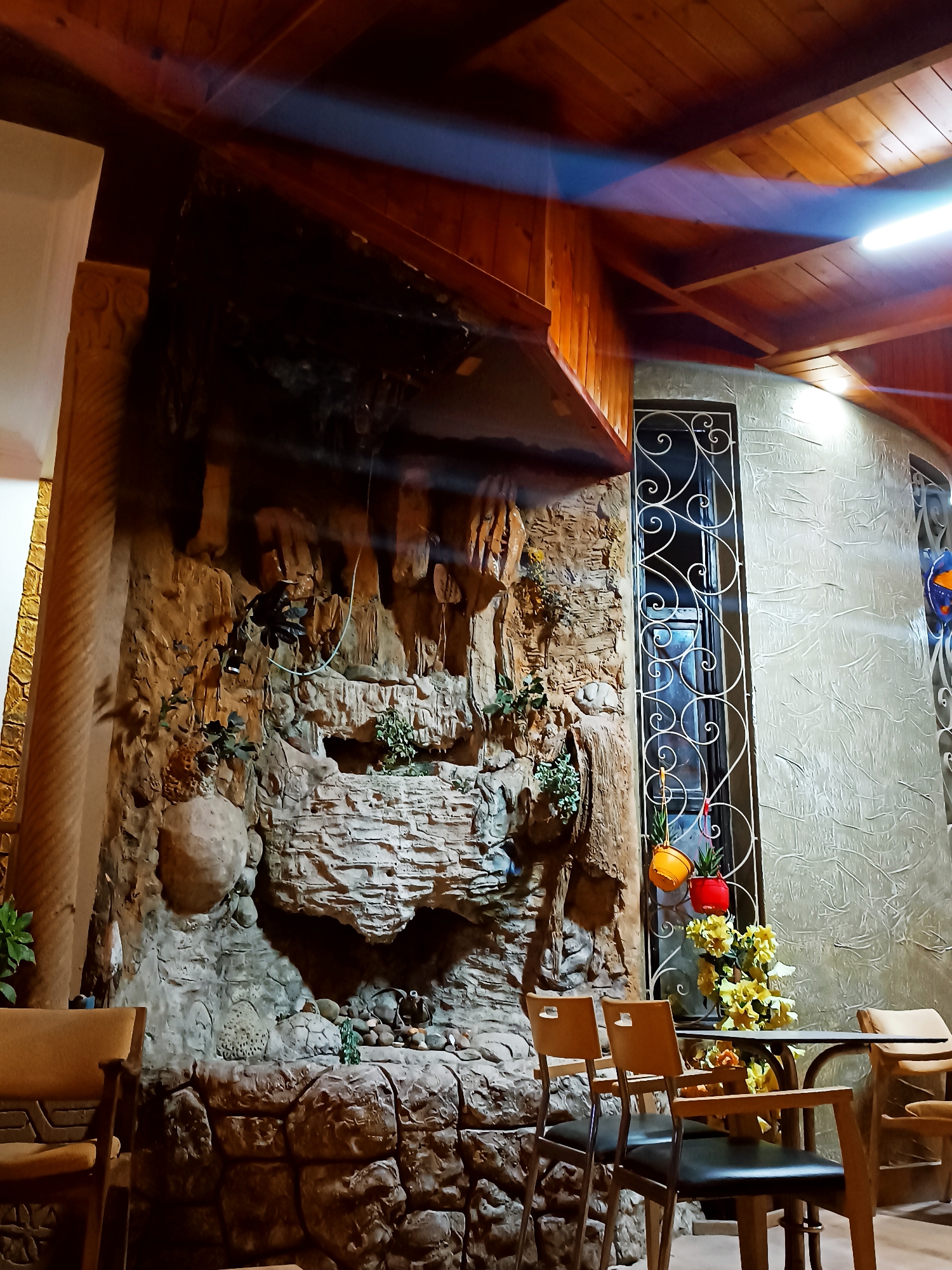 The image depicts an indoor seating area of a cafe or restaurant. The location might be Sidi Allal El Bahraoui, Morocco, although that detail isn't readily discernible from the visual information alone.
The focal point is a textured wall resembling a rock formation with a cascading waterfall effect. Various plants are integrated into the rock-like structure, adding a touch of nature. To the right of the rock formation is a decorative metal window frame with a white, ornate design.
In the foreground, there are several tables and chairs. The chairs appear to be made of wood with dark-colored seats. One table is adorned with a floral arrangement.
The ceiling has a wooden paneling design and a light fixture is illuminating the scene. The lighting suggests a warm and inviting atmosphere. The overall impression is of a themed interior, possibly intended to evoke a natural, rustic setting.