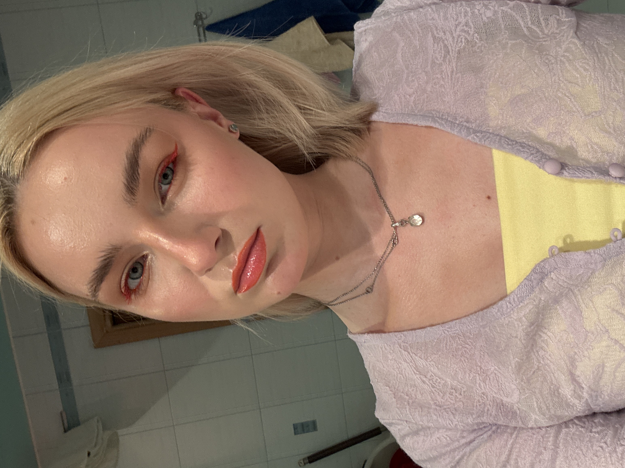 A young woman with blonde hair and fair skin is looking directly at the camera. She is wearing a light purple, textured cardigan over a yellow top, and a silver necklace with a pendant. Her makeup includes bright red eyeliner and a glossy coral lipstick. She is indoors, in what appears to be a bathroom with tiled walls.