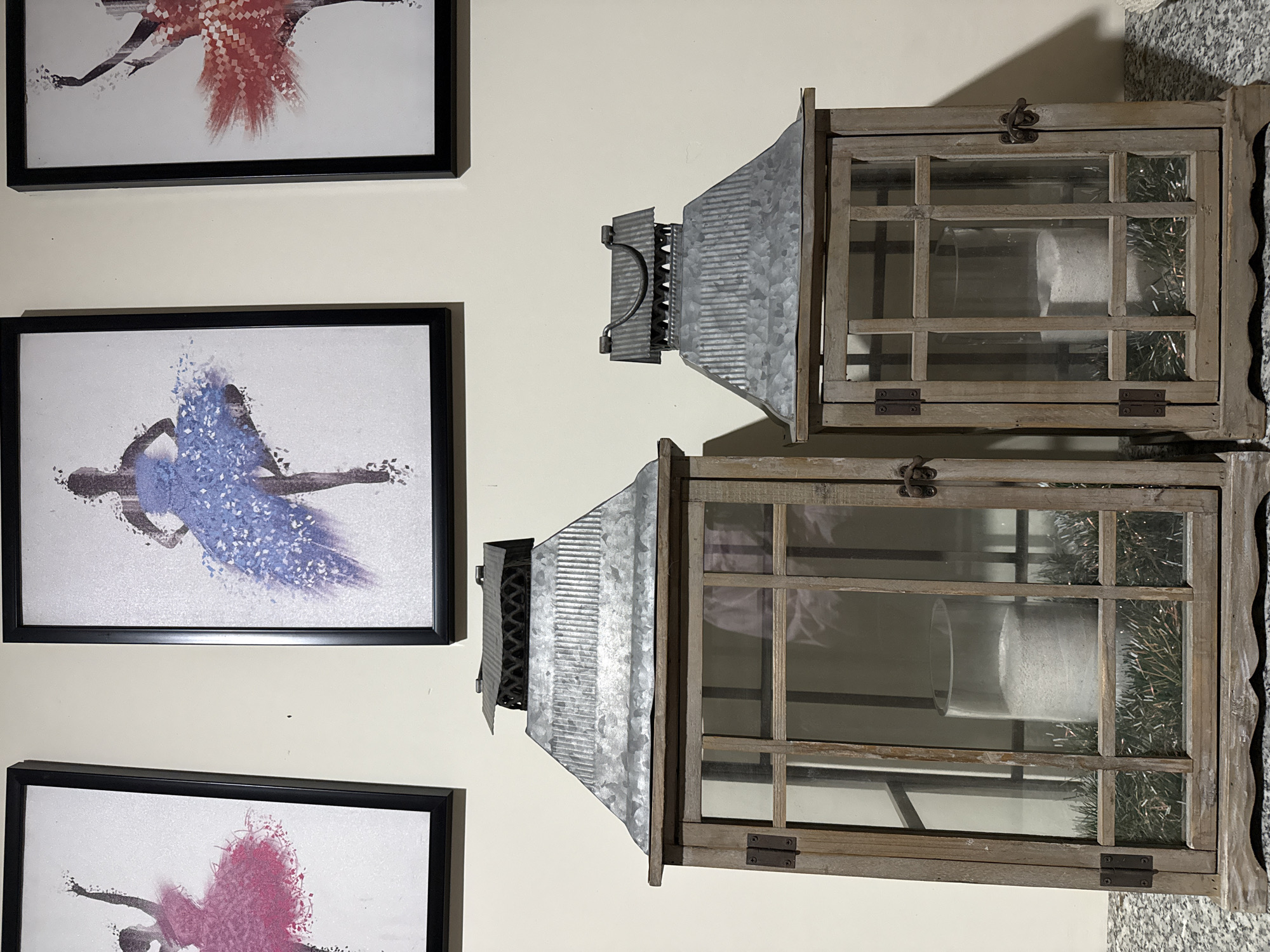 The media file displays a wall decorated with artwork and rustic lanterns. On the left side, there are three framed pictures featuring abstract depictions of dancers. The top picture shows a dancer in a red outfit, the middle picture a dancer in a blue tutu, and the bottom picture a dancer in a pink outfit. All dancers are rendered with a fragmented, pixelated style.
To the right of the artwork, two wooden lanterns are mounted on the wall. These lanterns have a rustic, farmhouse aesthetic, with distressed wooden frames and glass panes. Each lantern has a galvanized metal roof with a decorative, pointed shape. The upper lantern has a hinged latch at the top, and both lanterns appear to contain a white cylindrical object, possibly a candle or vase, and some greenery, likely tinsel or decorative foliage. The lighting suggests it is indoors, possibly in a residential setting, with warm light casting shadows on the wall. The overall impression is one of home decor with an artistic and vintage touch.