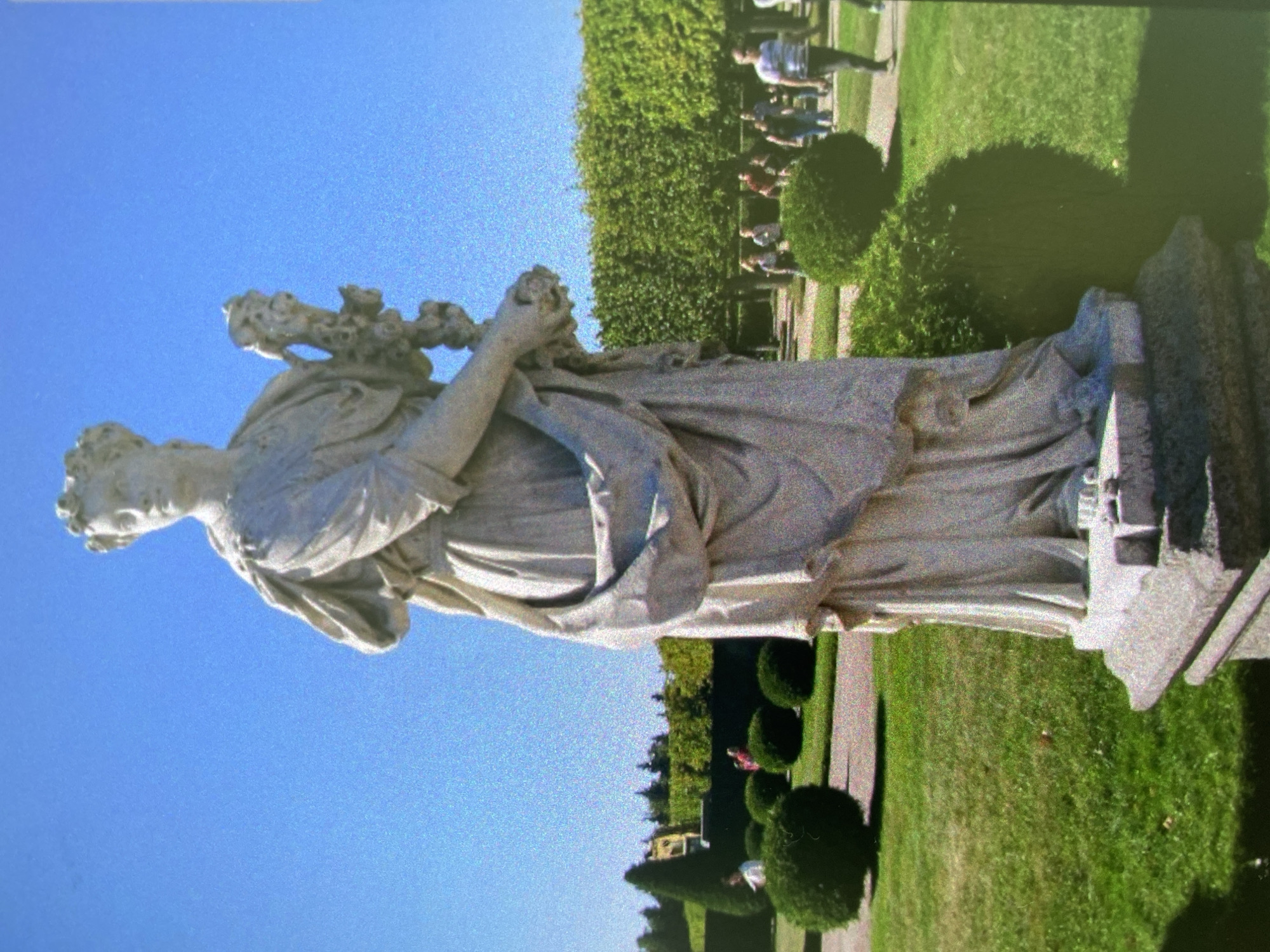 The image shows a white marble statue of a woman holding flowers in a garden setting. The statue is positioned on a stone base. The background consists of a bright blue sky and green landscape, including trimmed bushes and a patch of grass. In the distance, people are walking on a pathway. It depicts the Alexander Nevsky Lavra Cemetery in the city of Saint Petersburg, Russia.