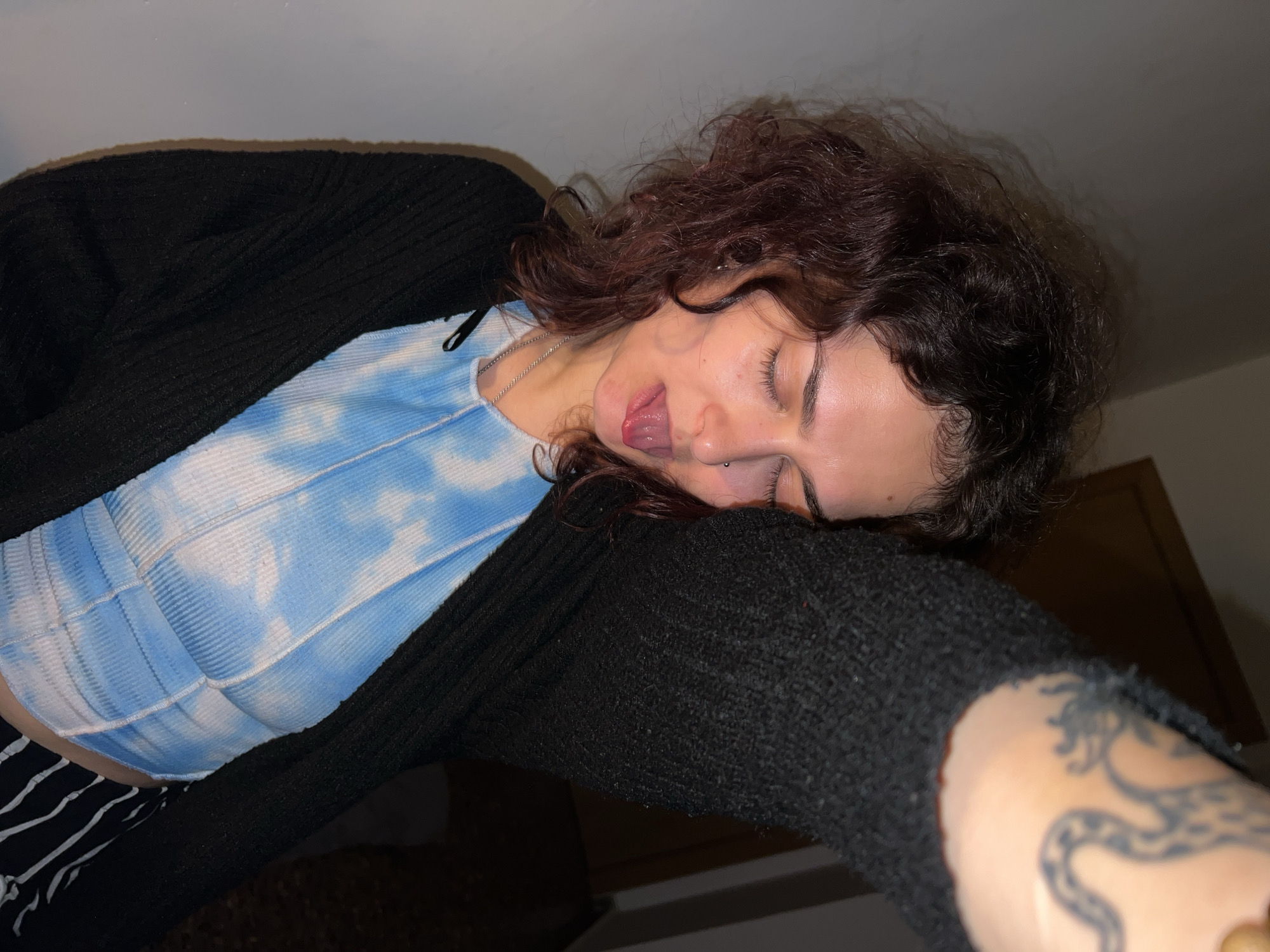 This close-up, horizontal image captures a young woman indoors in Picassent, Spain, in what appears to be an evening or nighttime setting due to the dim lighting. She is depicted lying down or leaning back, with her head tilted at an angle, eyes mostly closed, and tongue playfully sticking out.
The woman has curly, dark reddish-brown hair. She is wearing a ribbed, light blue and white tie-dye crop top, layered under a black ribbed cardigan or sweater. A silver chain necklace is visible around her neck. Distinctive features include an eyebrow piercing and a tongue piercing. On her left forearm, a dark, stylized tattoo, possibly depicting a snake or dragon, is partially visible. Her expression conveys a relaxed, perhaps whimsical or tired, yet lighthearted mood. The background is largely indistinct and dark, suggesting a plain wall or ceiling, with a hint of a wooden frame or furniture piece in the upper right. There is no visible text in the image.