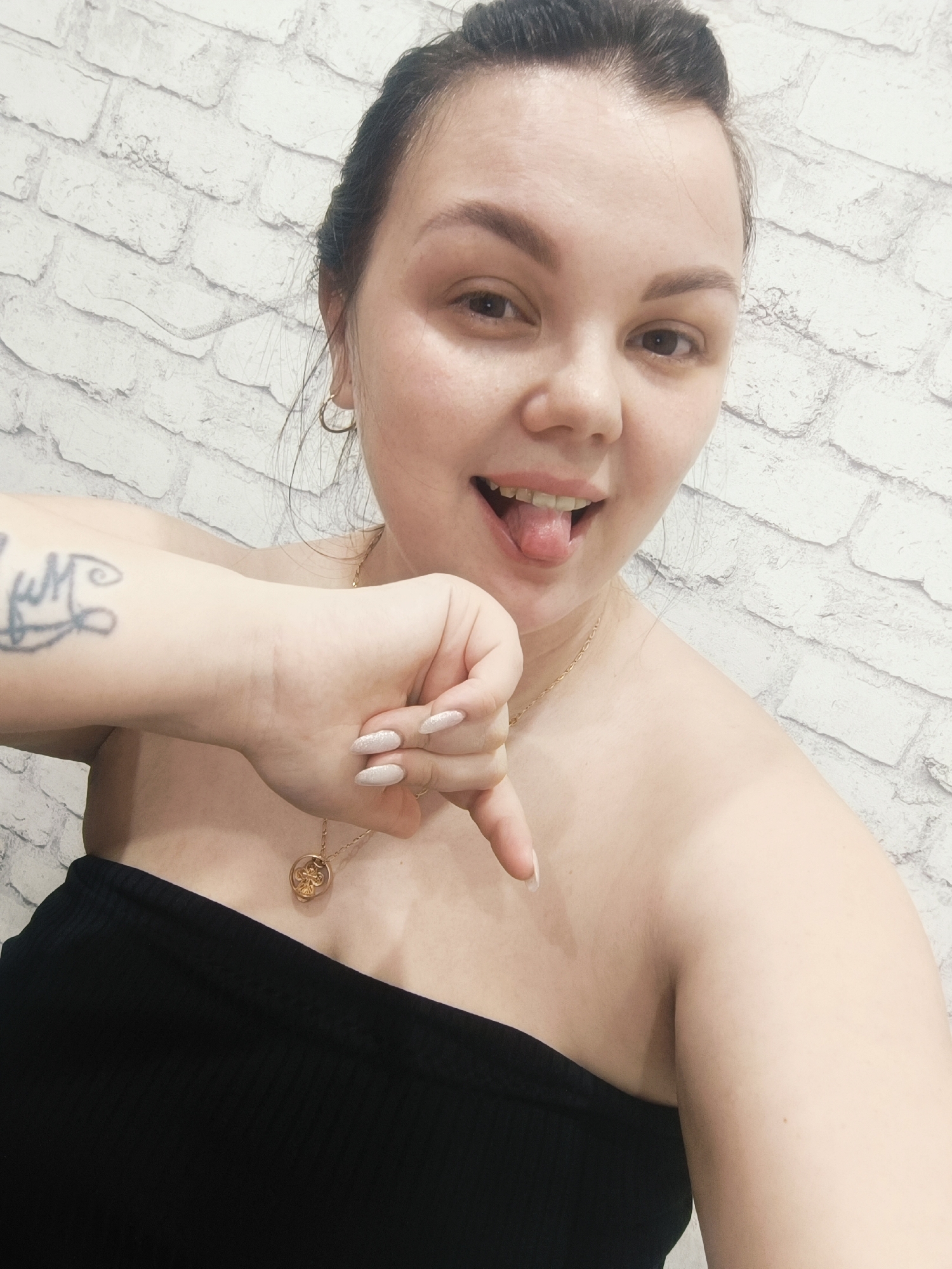 A medium shot captures a woman in front of a white brick wall. She has dark hair pulled back, fair skin, and is wearing a black tube top. She sticks her tongue out and gives a thumbs down gesture with her right hand, which has a tattoo on the wrist and manicured nails. She wears gold hoop earrings and a delicate gold necklace. The location is an unknown city and state, given the information.