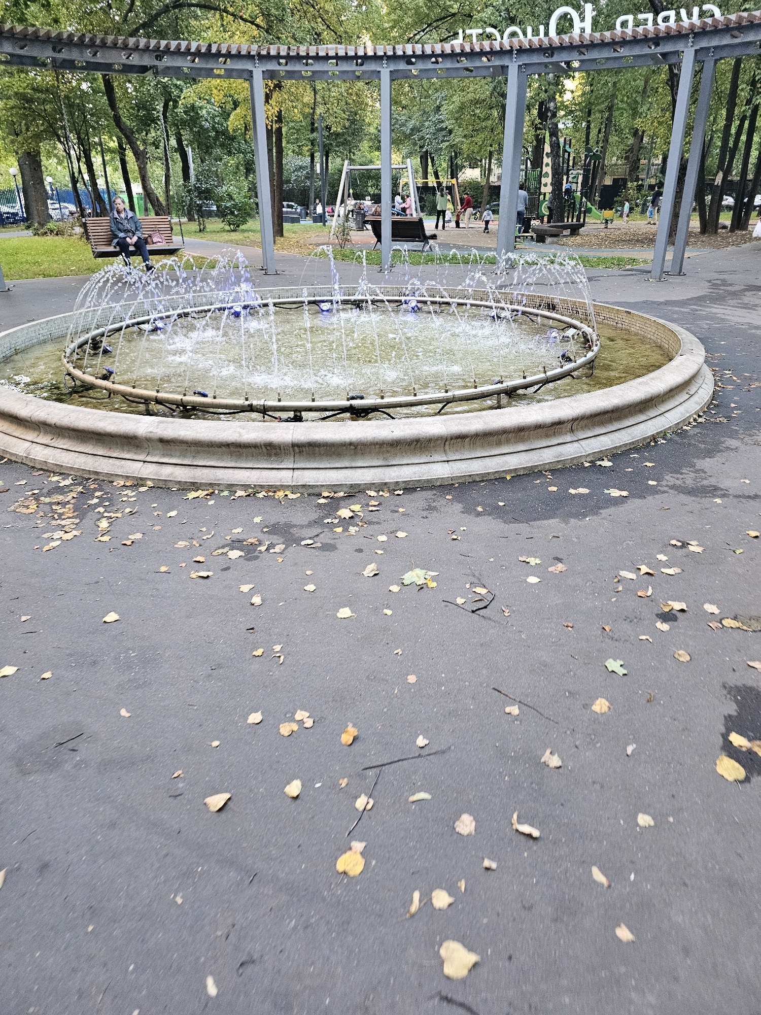 A circular fountain with illuminated water jets is in the foreground of an outdoor park. Fallen leaves are scattered across the wet asphalt path surrounding the fountain. In the background, a playground with swings and slides is visible, with several people of various ages interacting within the area. Behind the playground, trees with green and yellow foliage line the park, and a residential building is visible in the distance. The overall atmosphere suggests a public park in autumn, potentially in Khimki, Russia, given the Cyrillic inscription visible on the structure behind the fountain, which reads "КВЕДО 10" or similar. A woman is seated on a wooden bench to the left, facing the playground.