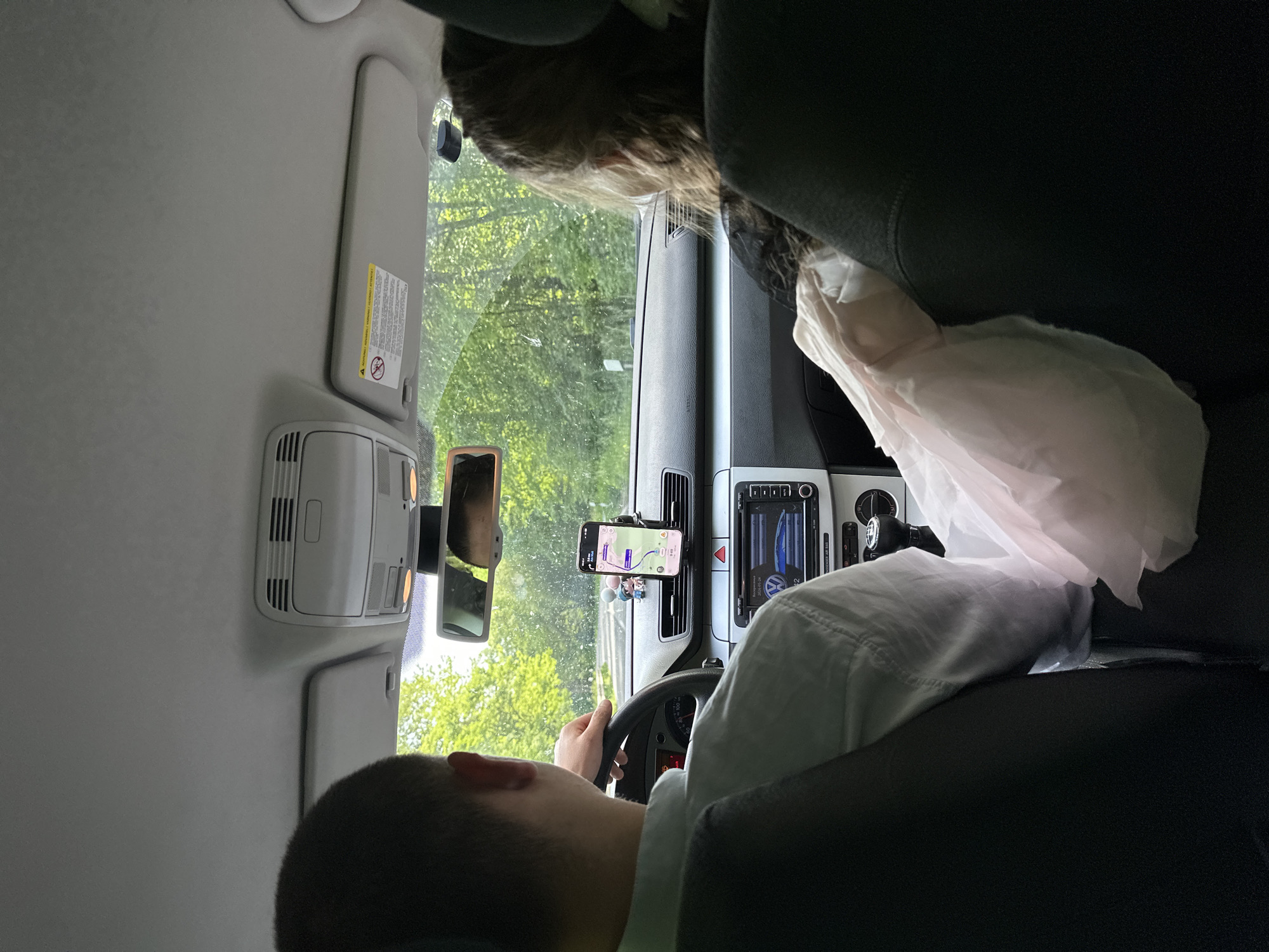 The image, likely taken from inside a car in Rechtenbach, Germany, shows a man driving. He is holding the steering wheel, with his right arm visible. A cell phone is mounted on the dashboard displaying a navigation application. The car's dashboard also includes a built-in display, and a plastic bag is positioned on the passenger seat. Outside the car, trees are visible through the windshield, suggesting a drive in a wooded or park-like area. A person with brown hair is sitting in the passenger seat. The car's interior lights and sun visor are visible overhead.
