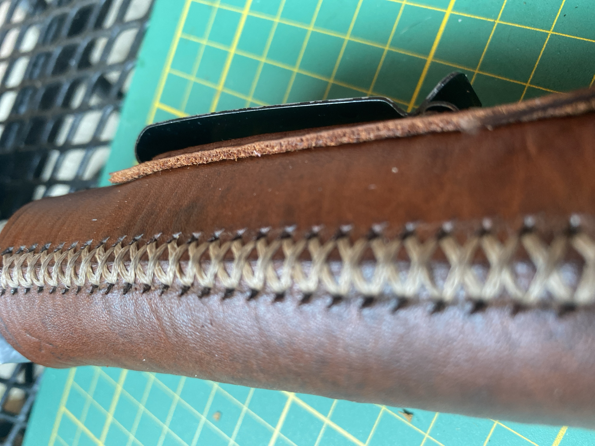 The image shows a close-up of a brown leather item, possibly a sheath or case, with decorative stitching. The stitching is a light tan color and forms a criss-cross pattern along the edge. The leather appears textured and slightly worn. Part of a black metal object is visible inserted into the leather item. The background consists of a green cutting mat with yellow grid lines, and a blurred black mesh structure. There are no people or animals visible in the image. The image does not provide any information that could be used to determine its location in the city of Lütjenwestedt, Germany.