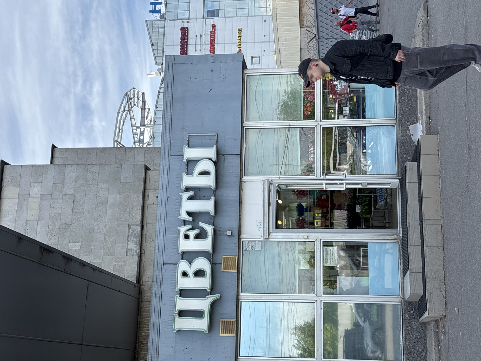 This is an urban street scene captured during the daytime, likely in Saint Petersburg, Russia. The primary subject is a building housing a shop, featuring a large sign and multiple glass-paned doors and windows on its facade. The prominent sign, in large white Cyrillic letters with a green outline, reads "ЦВЕТЫ" (TSVETY), which translates to "FLOWERS."
On the sidewalk in front of the shop, a young man stands, looking downwards. He is dressed in a dark denim jacket, dark pants, and a dark baseball cap. A red earbud is visible in his right ear. In the background, further down the street, two other individuals are visible: one wearing a white shirt and dark pants, and another in a red jacket.
The sky above is partly cloudy. In the upper left portion of the image, a large, intricate metallic structure, possibly decorative or part of a larger complex, is visible against the sky. The surrounding buildings are mostly commercial, with large glass windows and concrete or tiled facades.
Other visible text includes partial signs on a taller building in the background: "СМОРОД" (SMOROD) with smaller text below, possibly "товары для дома" (goods for home), "DOPADO.RU" (DORADO.RU), and another partial sign reading "ВКУСИЕ" (VKUSIE). Reflections of trees, the street, and a car are visible in the shop's glass windows. The ground consists of a paved sidewalk and asphalt street. The overall impression is a bustling urban environment.
