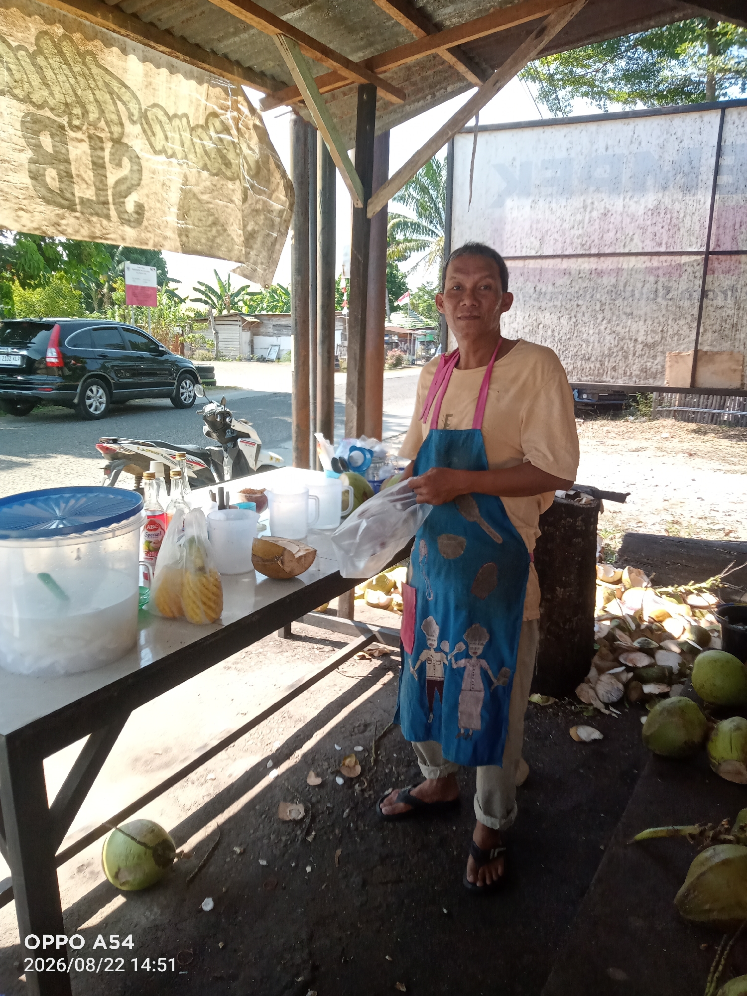 A medium-shot image shows a man standing at a street-side stall in Jambi, Indonesia, selling coconuts and drinks. The man is wearing a light-colored t-shirt, tan pants, and a blue apron adorned with cartoon figures. He is holding a plastic bag in his hands, and a large container filled with ice and possibly drinks is on the table to his left. Several coconuts, both whole and halved, are scattered around the stall, with some being prepared for sale. A black SUV and a motorcycle are parked nearby. The stall is covered by a corrugated metal roof supported by wooden beams, and a large banner with text is hanging above. The ground is dirt and debris, typical of an outdoor market setting. The overall impression is of a humble, open-air establishment.