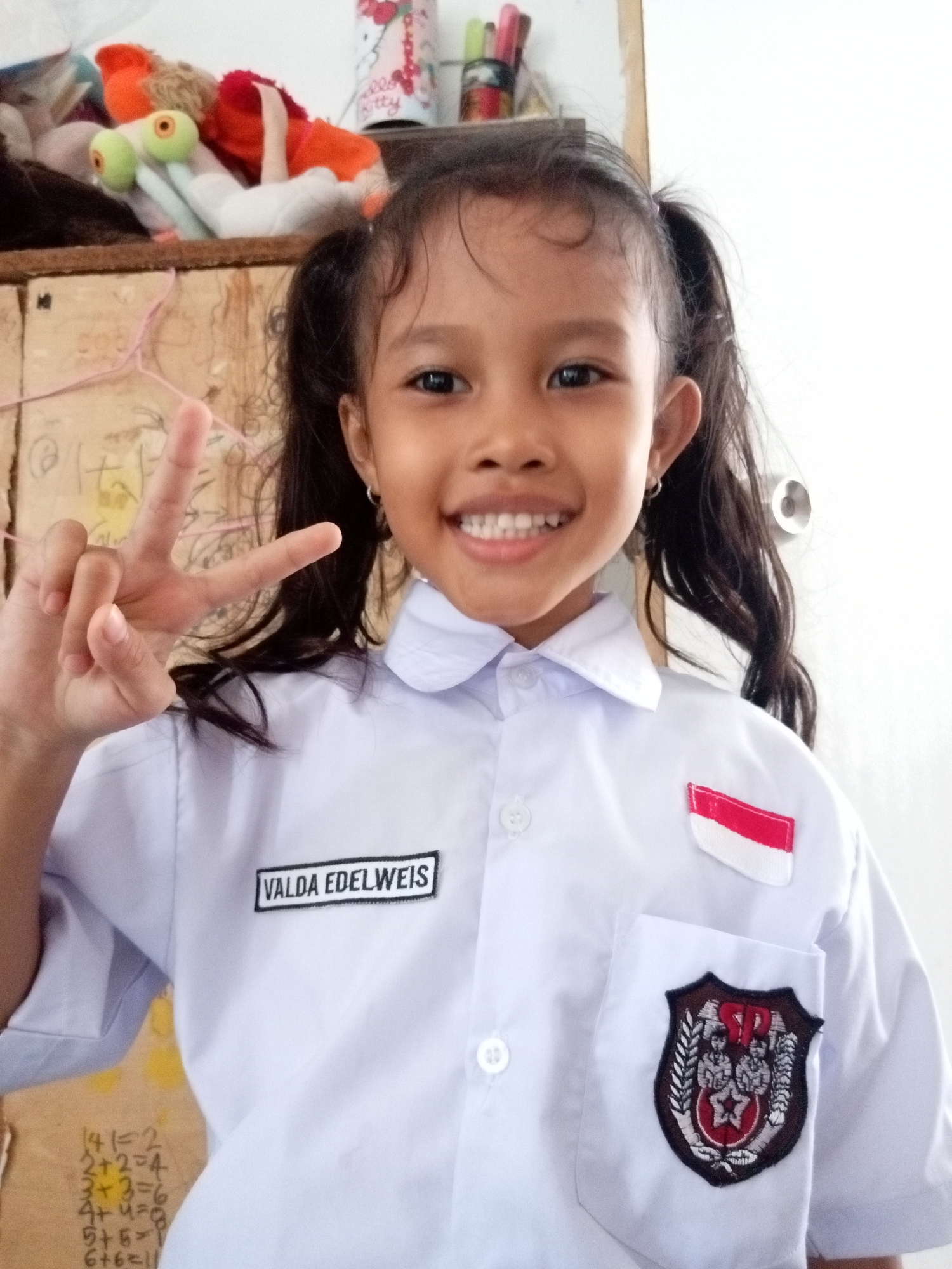 A young girl with dark hair pulled into two pigtails smiles at the camera, holding up a peace sign with her left hand. She is wearing a white school uniform with a name tag that reads "VALDA EDELWEIS" on the left side of her chest. A small Indonesian flag patch is visible on the right breast of her shirt, and a school emblem is on the left breast pocket. The background is blurry, but appears to be an indoor setting with a shelf containing stuffed animals and other items. The girl is likely in Batam, Indonesia, given the context of the uniform and flag.