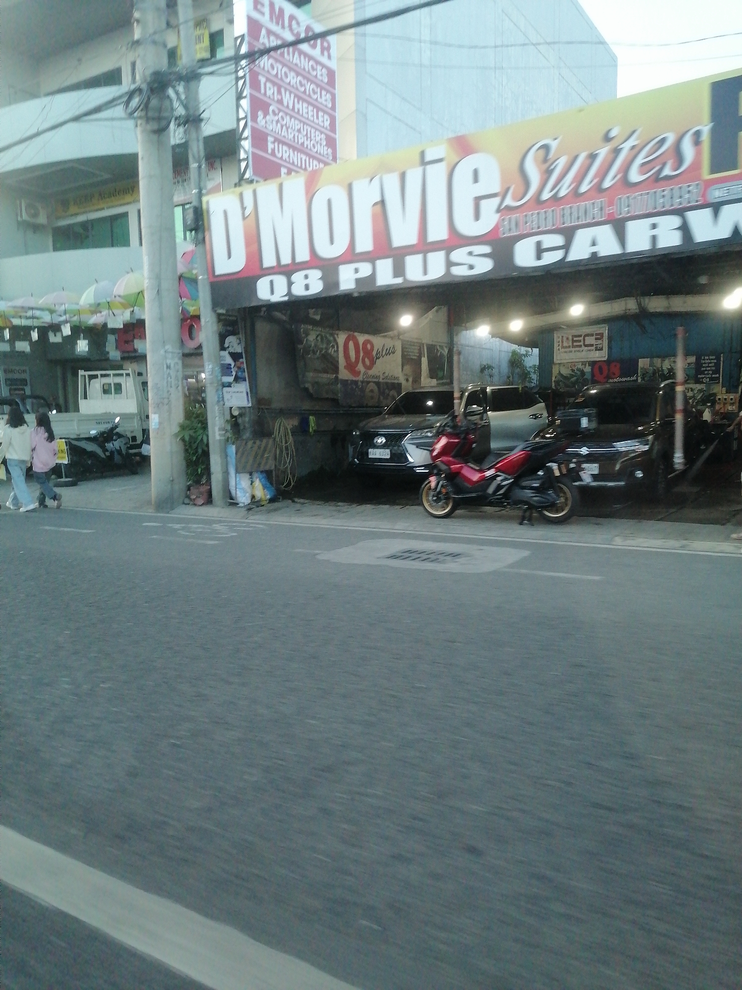 The image displays a street scene in Davao City, Philippines, specifically identifiable by the "D'MORVIE Suites SAN PEDRO BRANCH" sign. In the immediate foreground, a grey asphalt road with a partial white solid line on its left edge is visible.
On the right side of the frame, a red scooter with gold-colored wheels is parked in front of a commercial establishment identified as "D'MORVIE Suites 08 PLUS CARWASH," which features a prominent yellow and red sign. Inside this open-sided car wash facility, several vehicles are present, including a dark-colored SUV on the left, a silver pickup truck behind the scooter, and another dark-colored vehicle further right. The interior is illuminated by overhead lights, and cleaning equipment such as hoses and buckets are discernible.
In the mid-ground on the left, two women are walking from right to left on a paved area. The lead woman wears a light-colored long-sleeved top and blue jeans, while the woman behind her wears a pink long-sleeved top and dark pants. Both appear to be carrying items. A white light truck is parked nearby, positioned to the left of the car wash entrance.
Further into the background, several utility poles with a dense network of overhead wires are prominent. Behind these poles, a vertical red sign for "EMCOR" is visible on a building, listing products such as "APPLIANCES," "MOTORCYCLES," "TRI-WHEELER," "COMPUTERS & SMARTPHONES," and "FURNITURE." Adjacent to this, another building features "KEEP Academy" signage, with several decorative, light-colored umbrellas strung across an outdoor area in front of it. A larger, light-colored building with a modern facade is partially visible in the distant background. The overall lighting suggests either late afternoon, early evening, or an overcast day.