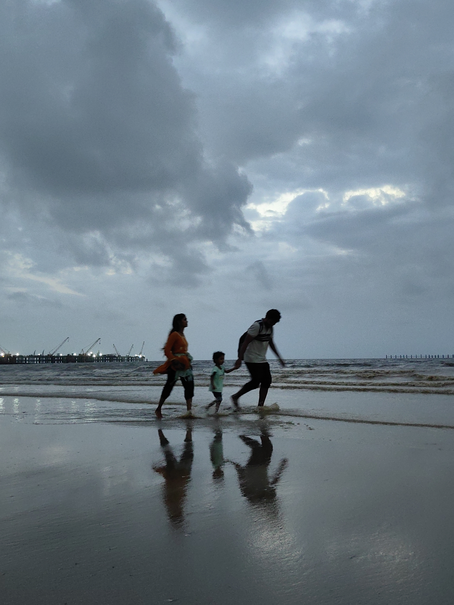 A family walks along the wet sand of a Mumbai beach, with the Arabian Sea in the background under a cloudy, overcast sky. The figures are silhouetted against the water and sky. In the foreground, the wet sand reflects the figures. In the middle ground, a man, a woman, and a young child walk from right to left. The man, wearing a t-shirt and shorts, carries the child on his shoulders. The woman, in a tunic and pants, walks beside them. In the background, a distant industrial complex with cranes is visible along the coastline.