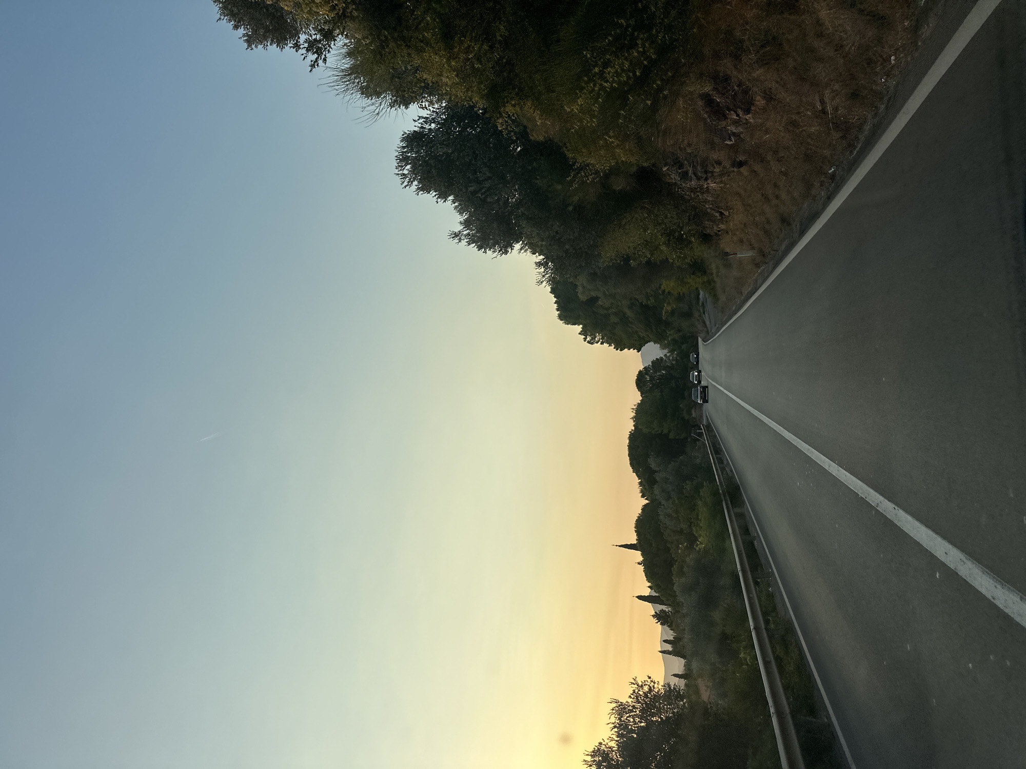 The image depicts a road winding through a landscape with trees and hills, under a pale sky transitioning from yellow to blue. The road is asphalt, with a white line marking the center. Several cars are visible on the road. On the left side, there's a metal guardrail and more trees and hills, with some structures that appear to be buildings and church spires in the distance. On the right side, the road is bordered by a rocky embankment with dense vegetation. The time of day appears to be late afternoon or early evening, given the warm hues in the sky. The location is identified as Sveti Stefan, Montenegro.