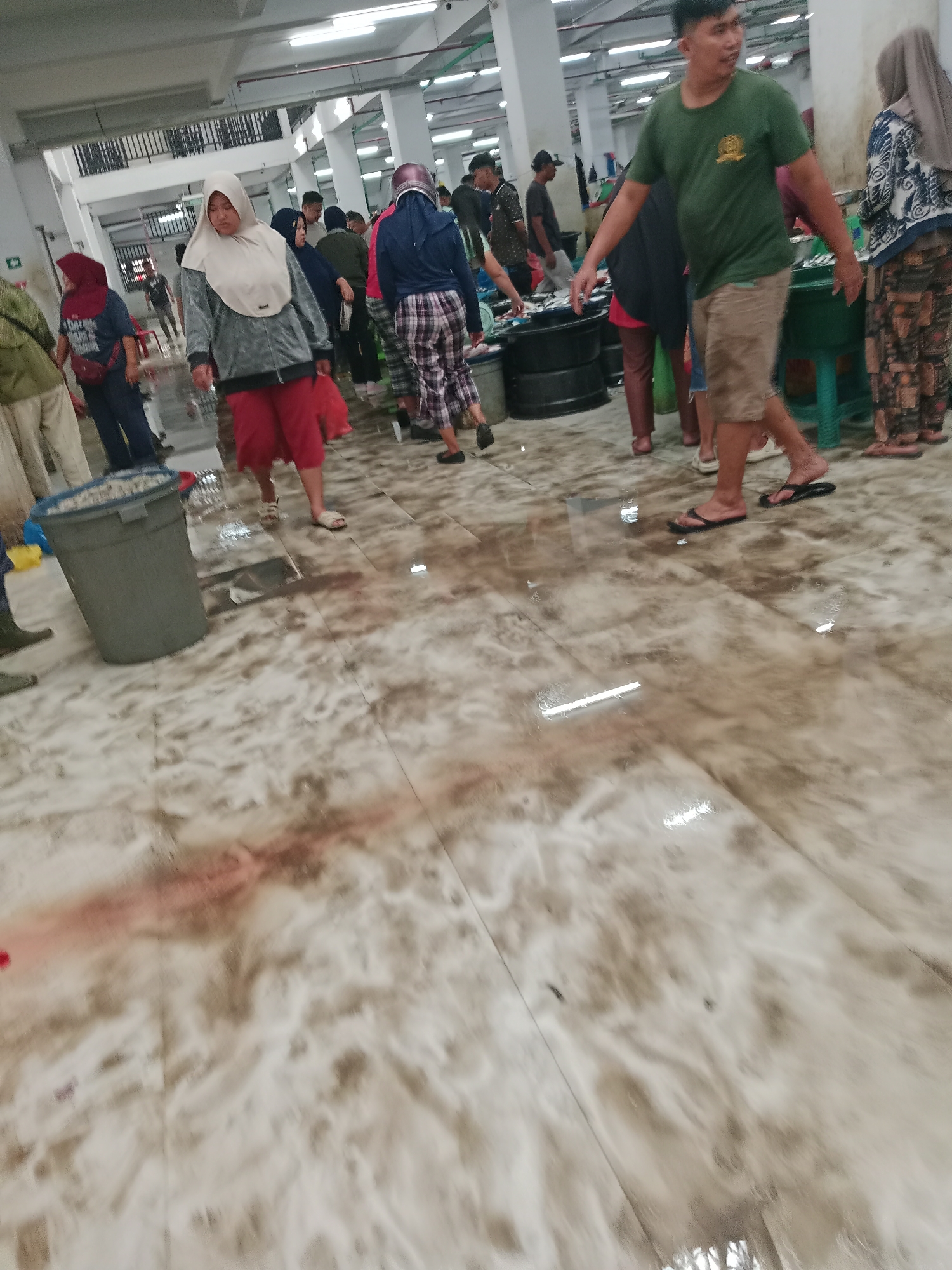The image depicts an indoor market scene in the City of Binjai, Indonesia. The floor is wet and appears to be a tiled surface with a marbled or polished finish, reflecting the overhead lighting. There are several individuals visible, navigating the market.
In the foreground, a person wearing a green t-shirt and khaki shorts is walking to the right. Behind them, a woman in a headscarf and patterned clothing is also walking. To the left, a large grey trash bin is positioned on the wet floor.
Further into the background, more people are dispersed throughout the market. Some are carrying bags or baskets, suggesting they are shopping or working. There are also large, dark circular containers, possibly for holding goods or water. The environment appears to be a covered market space with fluorescent lighting fixtures visible on the ceiling. The overall impression is of a busy, functional marketplace with some wetness on the floor, common in such environments.