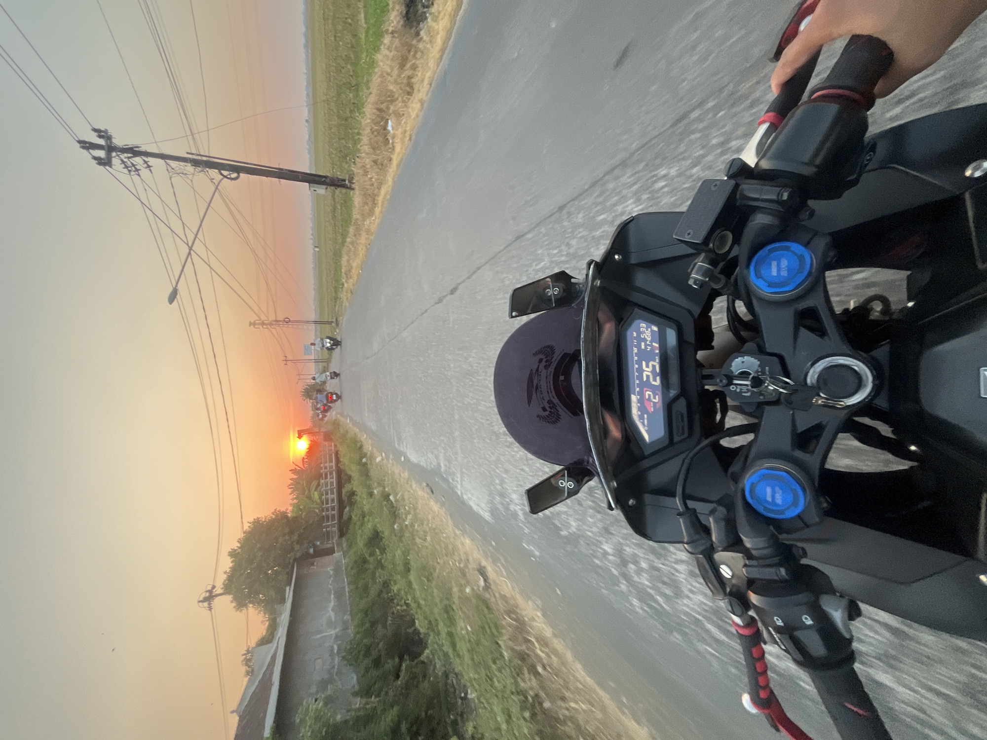 The image shows a point-of-view shot from a motorcycle on a paved road at sunset. The sky is a warm gradient of orange and yellow, with the sun low on the horizon. In the foreground, the handlebars and dashboard of the motorcycle are visible, with a digital display showing "42 25" and "5:33 41H68". To the left of the road, there is a building with a gray roof and vegetation. A small bridge crosses a waterway. Further down the road, several other motorcycles and a few people are visible. To the right of the road, there is a field of dry grass and green crops. Utility poles with numerous wires are prominent on the right side of the frame. The overall impression is of a rural scene in Sendang, Indonesia, at dusk.