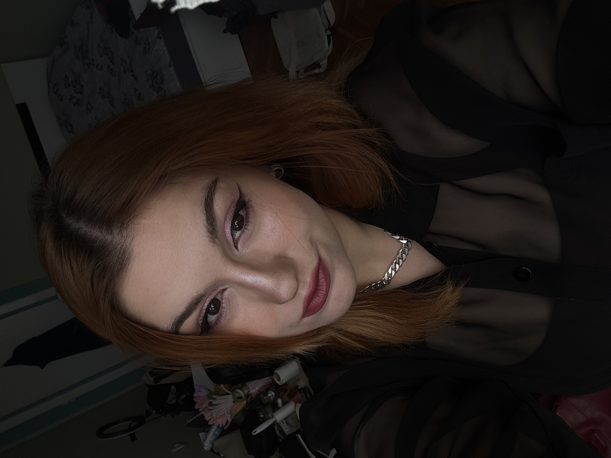 A close-up, slightly low-angle selfie of a young woman with reddish-brown hair. She has light skin, pink eyeshadow, winged eyeliner, and dark red lipstick. She is wearing a sheer black button-down shirt and a silver chain necklace. The background is a cluttered interior, with a bed visible, including pillows and a comforter. Other items such as what appear to be household goods, a ring light, and artificial flowers are also visible. The image is not readily identifiable in terms of a specific location within Varna, Bulgaria, due to the indoor setting and lack of distinguishable landmarks.