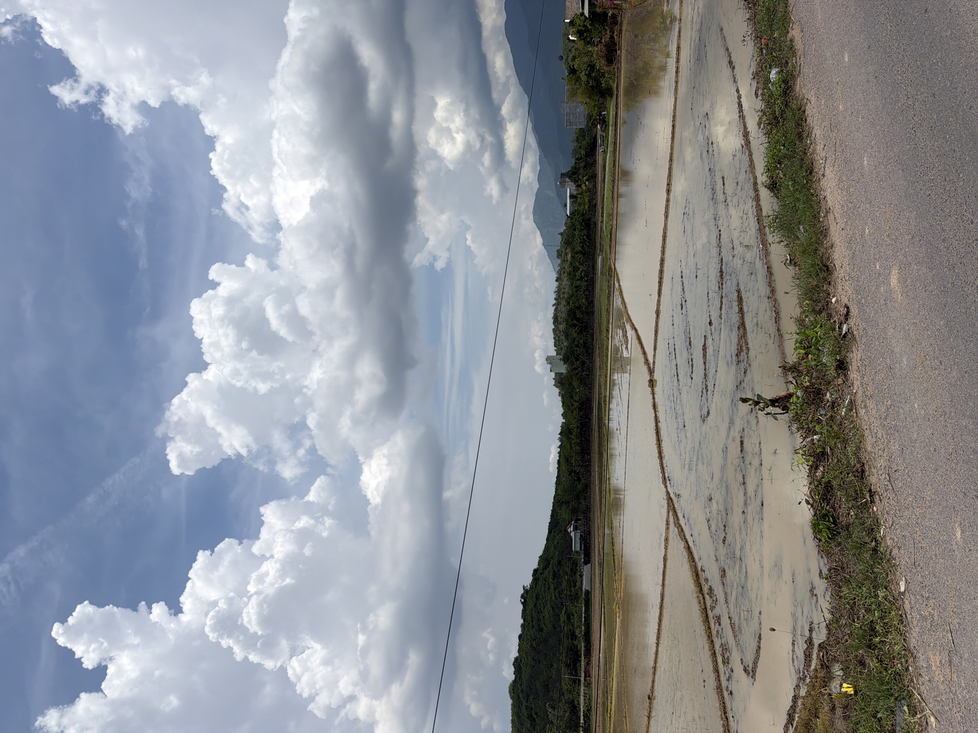 The image captures a wide view of flooded rice paddies in Diên Thọ, Vietnam, under a dramatic sky. The foreground shows a portion of an asphalt road and a grassy verge, with some scattered litter. The middle ground is dominated by meticulously sectioned rice paddies filled with murky water, reflecting the sky. Some raised earthen borders divide the paddies, with faint patterns of mud and possibly young plants visible within them. In the background, a densely forested hill with a small red-roofed building on its slope forms a backdrop, transitioning to rolling hills and distant mountains that fade into the hazy horizon. A prominent dark electrical wire stretches horizontally across the upper half of the image, bisecting the sky. The sky itself is a dynamic mix of bright white, puffy cumulus clouds against patches of blue, and darker, more overcast gray clouds that suggest recent or approaching rain. No people or animals are visible in the frame.