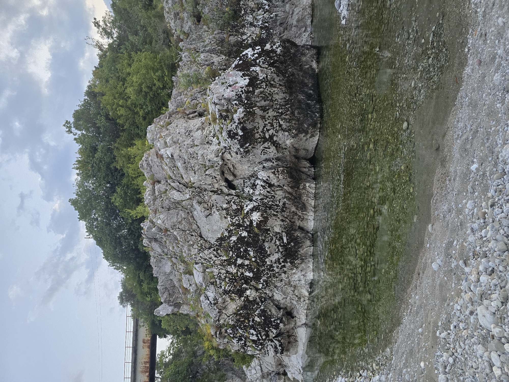 The image depicts a natural scene of a rocky riverbed and surrounding vegetation, with a bridge visible in the distance. The foreground shows a shallow river with clear water revealing numerous smooth pebbles on the riverbed. The riverbank is composed of grey and white pebbles and sand.
Dominating the mid-ground is a large, textured rock formation with patches of dark, moss-like growth. This rock face is partially submerged in the river. Behind the rock formation, lush green trees and foliage form a dense backdrop.
In the upper left portion of the image, a rusty metal bridge with red railings spans across the scene, partially obscured by trees. The sky is overcast with grey clouds.
The location is identified as Bioče, Montenegro, near the Morača River, which is known for its picturesque gorges and natural beauty. The scene captures a tranquil, wild environment with elements of man-made infrastructure (the bridge) integrated into the landscape. No people or animals are visible in the image.
