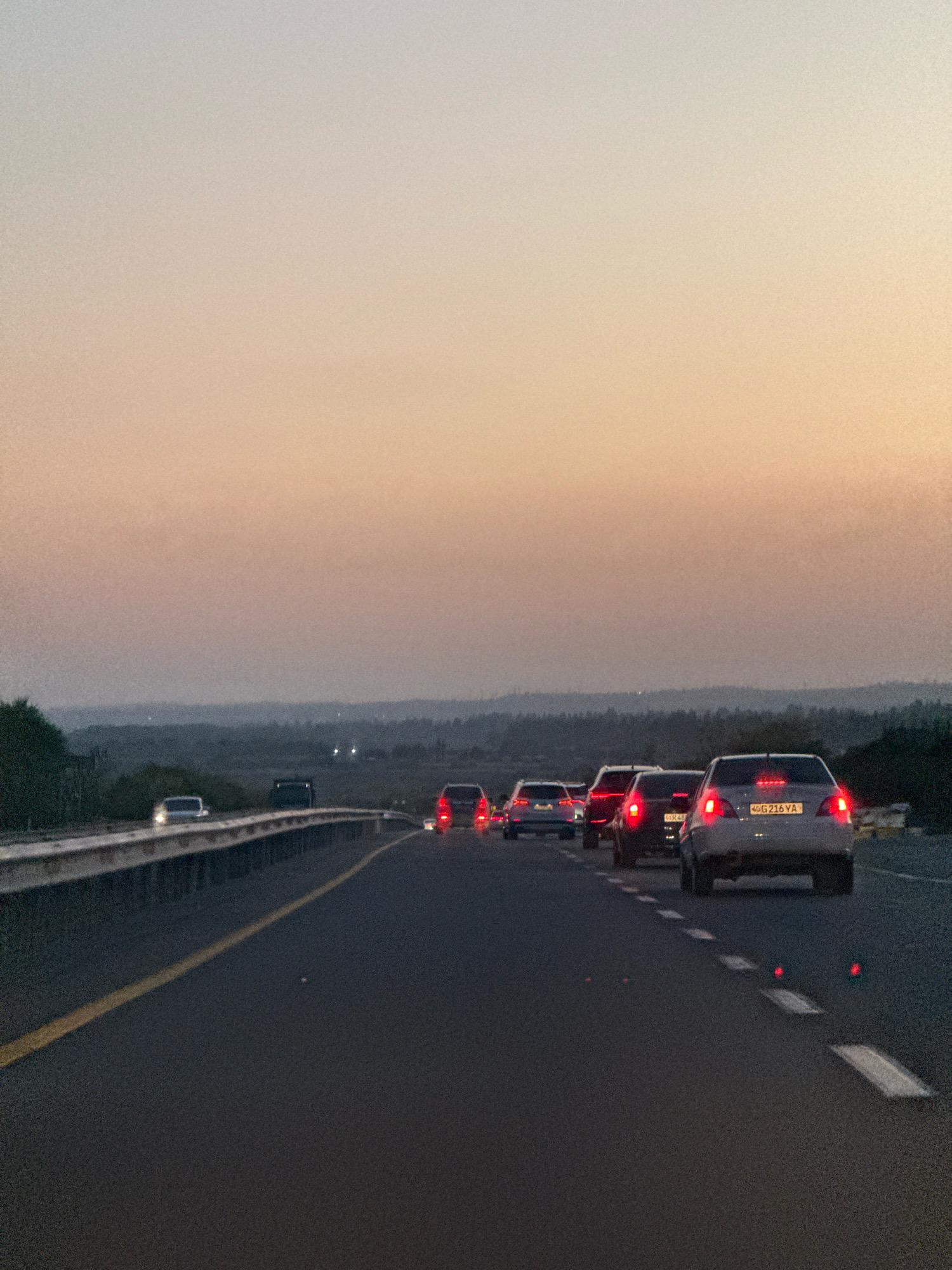 A view from the perspective of a moving vehicle on a highway at dusk. Multiple cars are traveling in the same direction, forming a queue. The sky displays a gradient of colors, transitioning from a light peach hue near the horizon to a pale blue at the top. The landscape in the distance consists of rolling hills with a light dusting of trees. The road surface is asphalt, marked with a solid yellow line on the left and broken white lines on the right. In the foreground, the rear of a silver sedan with license plate "40G 216 YA" is visible, its brake lights illuminated. Other vehicles with their brake lights on are also present in the traffic jam. The environment appears to be an open area outside of a dense urban center. There are no people clearly visible. This scene is consistent with a highway route approaching or leaving the city of Dumalak, Uzbekistan, during evening traffic.