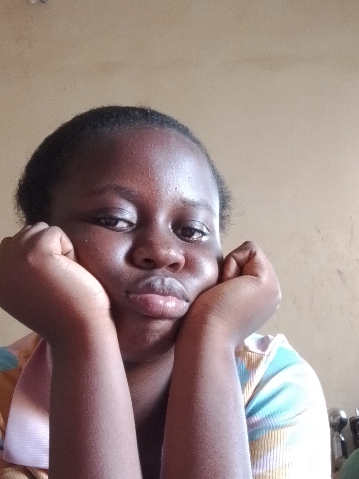 An image taken in OGBOKO, Nigeria, depicts a close-up portrait of one individual, a young female, positioned centrally. Her dark hair is pulled back, revealing her forehead. She has dark skin and is making a pouty expression with slightly parted, glossy lips. Her chin rests on her cupped hands, with her forearms angled upwards. She is wearing a multi-colored, ribbed shirt featuring horizontal stripes or patterns in light blue, white, and beige tones. The background consists of a plain, light brown wall. On the far right, in the blurred background, several indistinct metallic objects are partially visible, suggesting a domestic indoor setting.