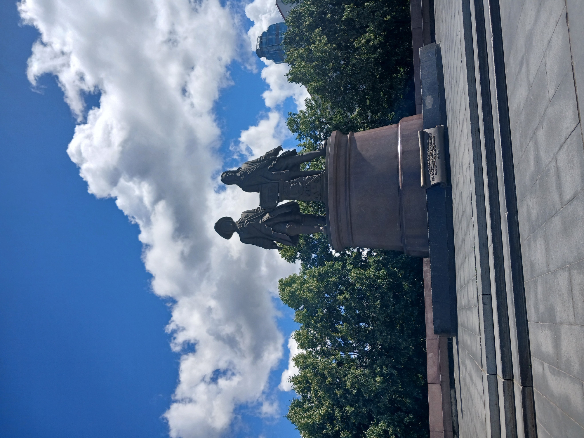 A bronze statue of two men stands on a cylindrical pedestal in a paved plaza. The statue depicts two figures in historical attire. The man on the left wears a hat and a coat, while the man on the right has long hair and also wears a coat. They are positioned side-by-side, with the man on the right appearing to hold a book or scroll. A bronze plaque with text is affixed to the front of the pedestal.
Behind the statue, a dense line of lush green trees forms a backdrop. Beyond the trees, a modern blue-glass skyscraper rises into a bright blue sky filled with fluffy white clouds. The plaza is paved with large square tiles and features several wide, shallow concrete steps leading up towards the monument. The scene suggests an outdoor public space, likely a park or square, in a city setting. This is the Monument to Vasily Tatishchev and Afanasiy Gerasimov, located in Yekaterinburg, Russia.