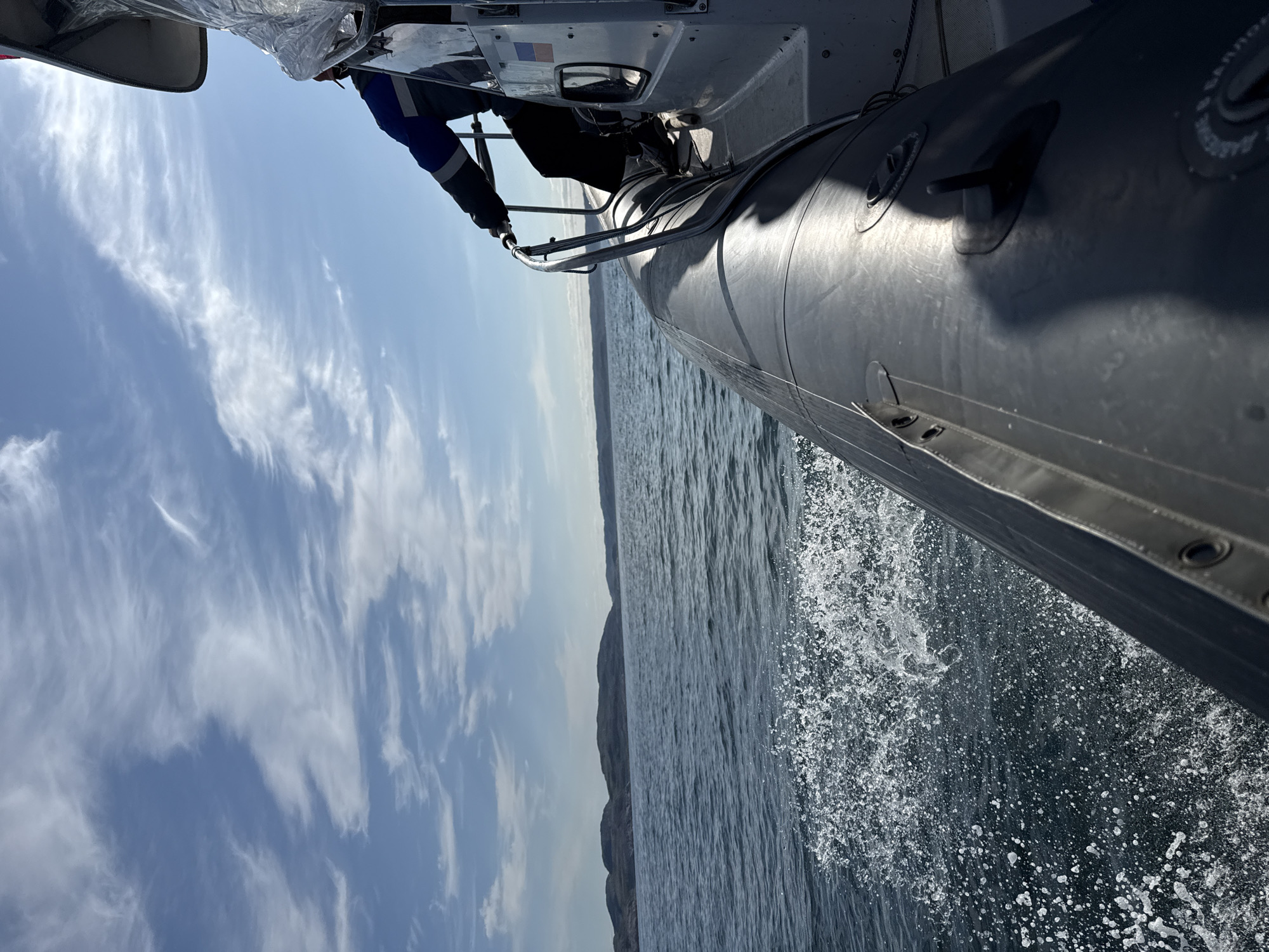 A person wearing a blue and black jacket with reflective stripes and black pants is holding onto the railing of a rigid-hulled inflatable boat (RHIB) while it moves through choppy, dark blue water. The boat's dark gray inflatable tube is visible in the foreground, with white spray splashing from its wake. In the background, a rugged, hilly coastline under a partly cloudy blue sky is visible. A small Russian flag is attached to the side of the boat. The image was taken from the perspective of someone on the boat, looking forward and to the side. The scene likely depicts travel on the coast of Teriberka, Russia, a location known for its Arctic environment and coastline.