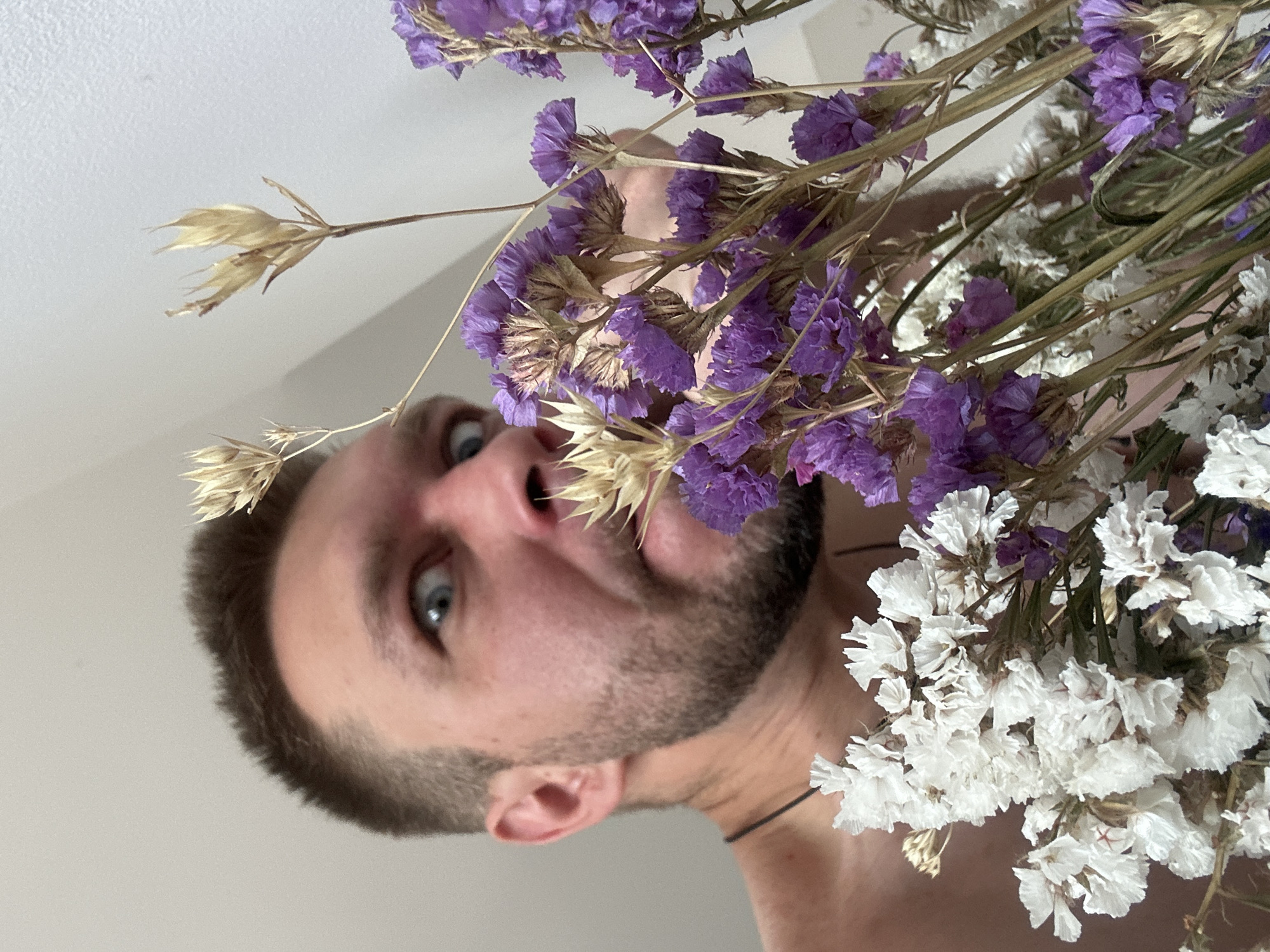 The media file is a close-up, upside-down photograph of a man's face emerging from a bouquet of dried flowers. The man has short, dark hair and stubble on his face. His eyes are wide and looking directly at the camera, conveying surprise or curiosity. The bouquet consists of clusters of small purple and white flowers, along with some dried wheat-like stalks. The background is a plain, light-colored surface, possibly a wall or ceiling. The lighting suggests an indoor setting, possibly during the daytime. The image is a selfie, taken from a low angle, with the man's face framed by the flowers. There is no visible text in the image. The overall impression is playful and slightly unconventional.