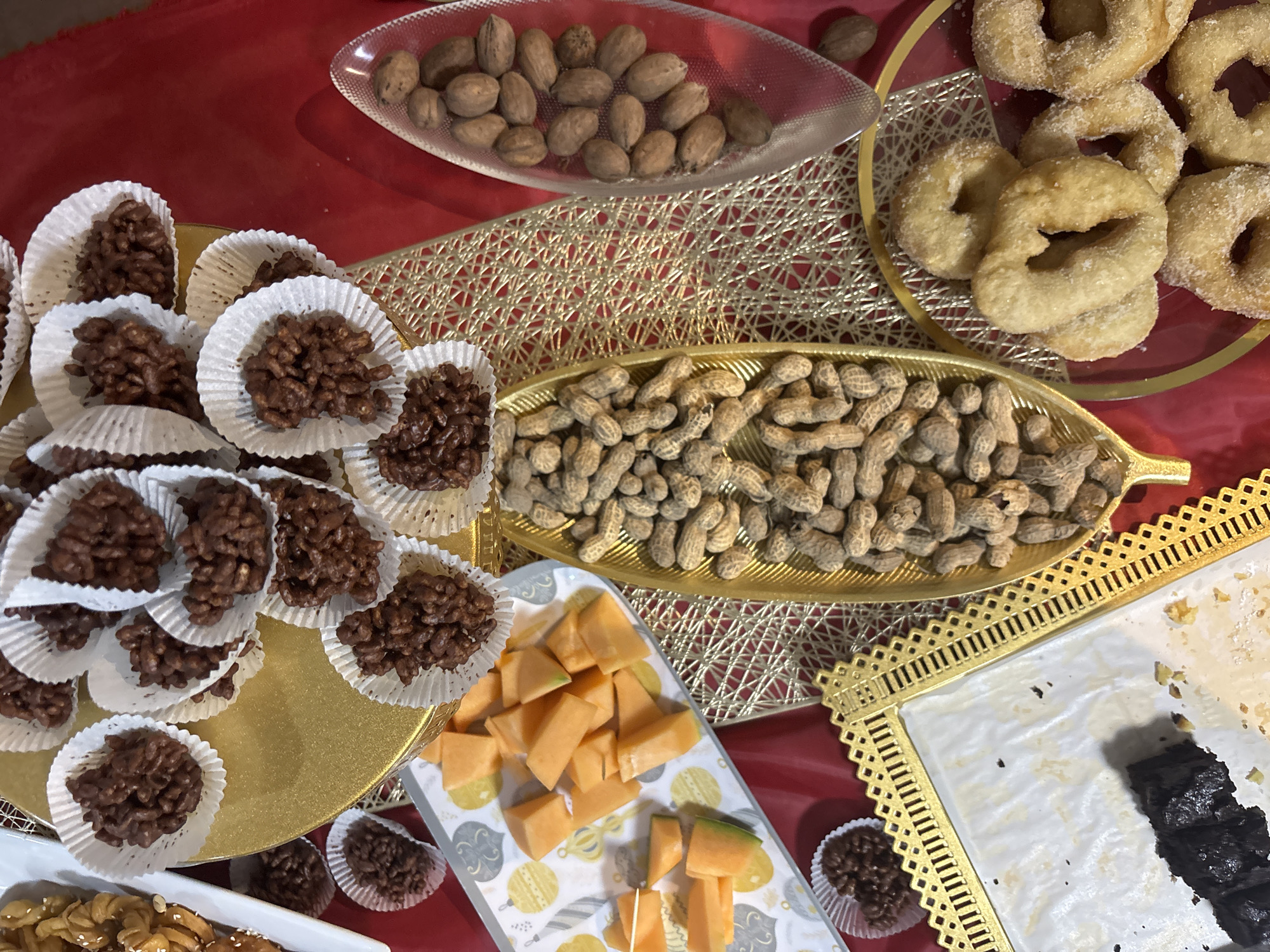 The image shows an assortment of snacks and desserts arranged on a red surface, likely a tablecloth. The scene is indoors, presumably a home or event space.
The food items include:
- Small chocolate clusters in white cupcake liners, densely arranged on a gold tray.
- Peanuts in their shells, presented in a gold, leaf-shaped dish.
- Fried dough pastries, dusted with sugar, on a red and gold tray.
- Shelled nuts in a clear glass dish.
- Cubed cantaloupe on a patterned plate.
- Remnants of what appear to be chocolate brownies on a gold-trimmed white tray.
Given the absence of people and contextual clues, determining the specific location within Huixquilucan de Degollado, Mexico is not possible. The scene represents a dessert or snack spread, likely prepared for a gathering or celebration.