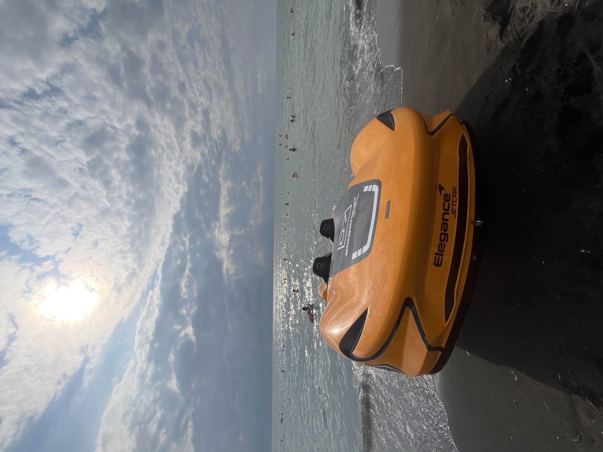 This image depicts an orange and black "Elegance JETCAR" parked on a dark, sandy beach in Ureki, Georgia. The jetcar is positioned facing slightly left, with its front end closest to the viewer and partially submerged in shallow water where small waves are breaking. In the background, the Black Sea stretches out, populated by numerous people swimming and bathing. The sun is visible in the sky, partially obscured by scattered clouds, casting a bright glare on the water. The overall scene suggests a warm, sunny day at a beach resort.