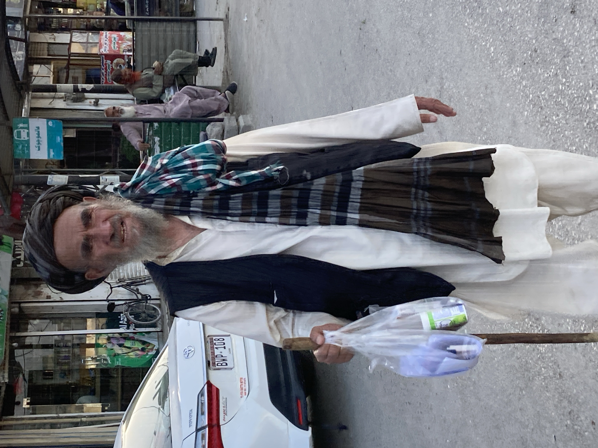 An elderly man stands on a street in Quetta City Tehsil, Pakistan. He wears a traditional turban, vest, and shawl. He holds a plastic bag containing items and leans on a wooden cane. Behind him are parked cars, storefronts, and two men reclining on chairs near a building.