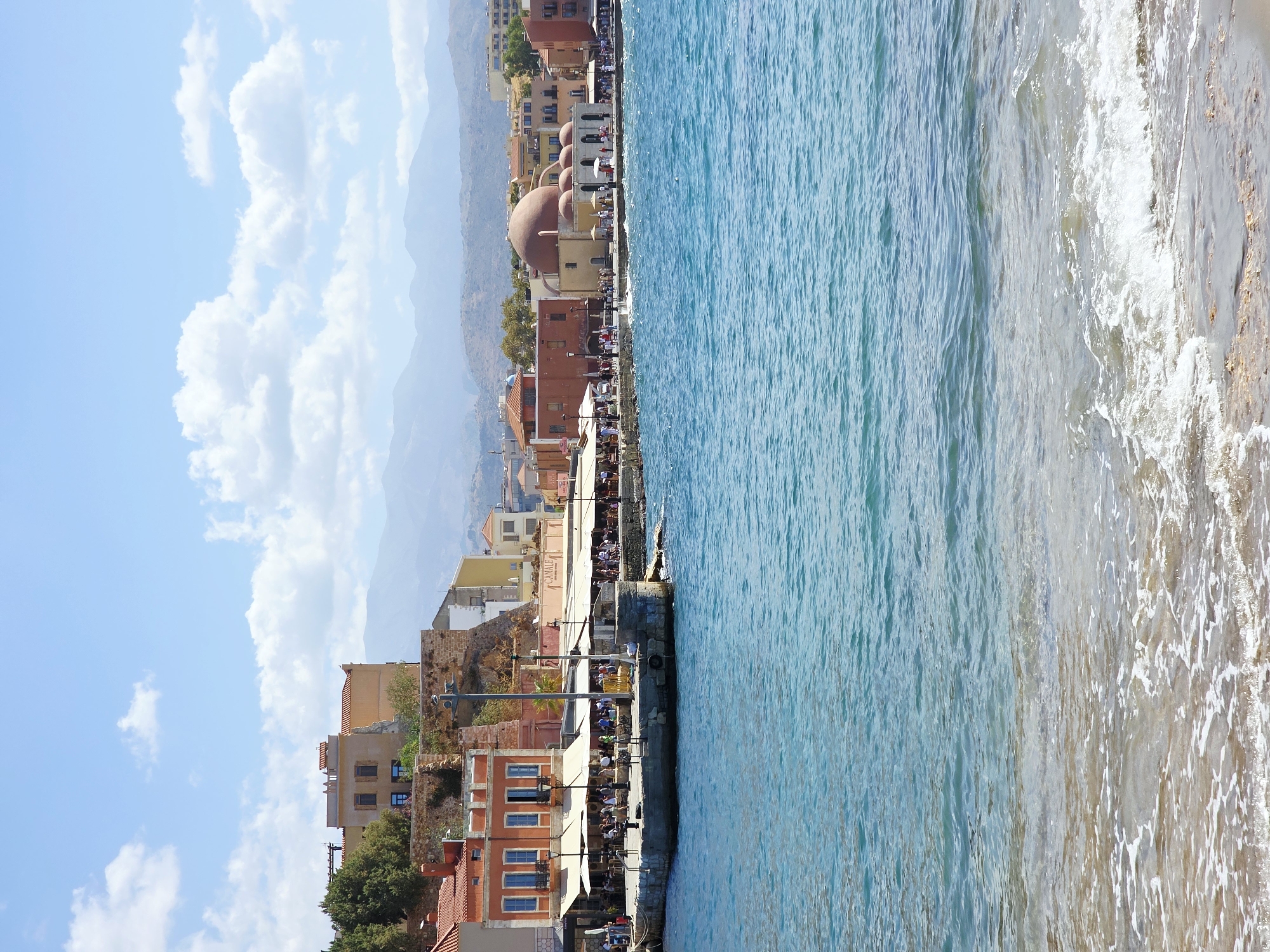 The image shows a sunny day at the harbor of Chania, Crete, Greece. In the foreground, the turquoise water of the sea laps gently onto a sandy and pebbly beach. A breakwater extends into the water, lined with buildings that house restaurants and cafes. Many people are seated at tables, some under umbrellas, enjoying the seaside atmosphere. The buildings are a mix of colors, with a prominent orange building on the left and a building with a large dome in the center-right. Beyond the harbor, a range of mountains is visible in the hazy distance under a bright blue sky dotted with white clouds.