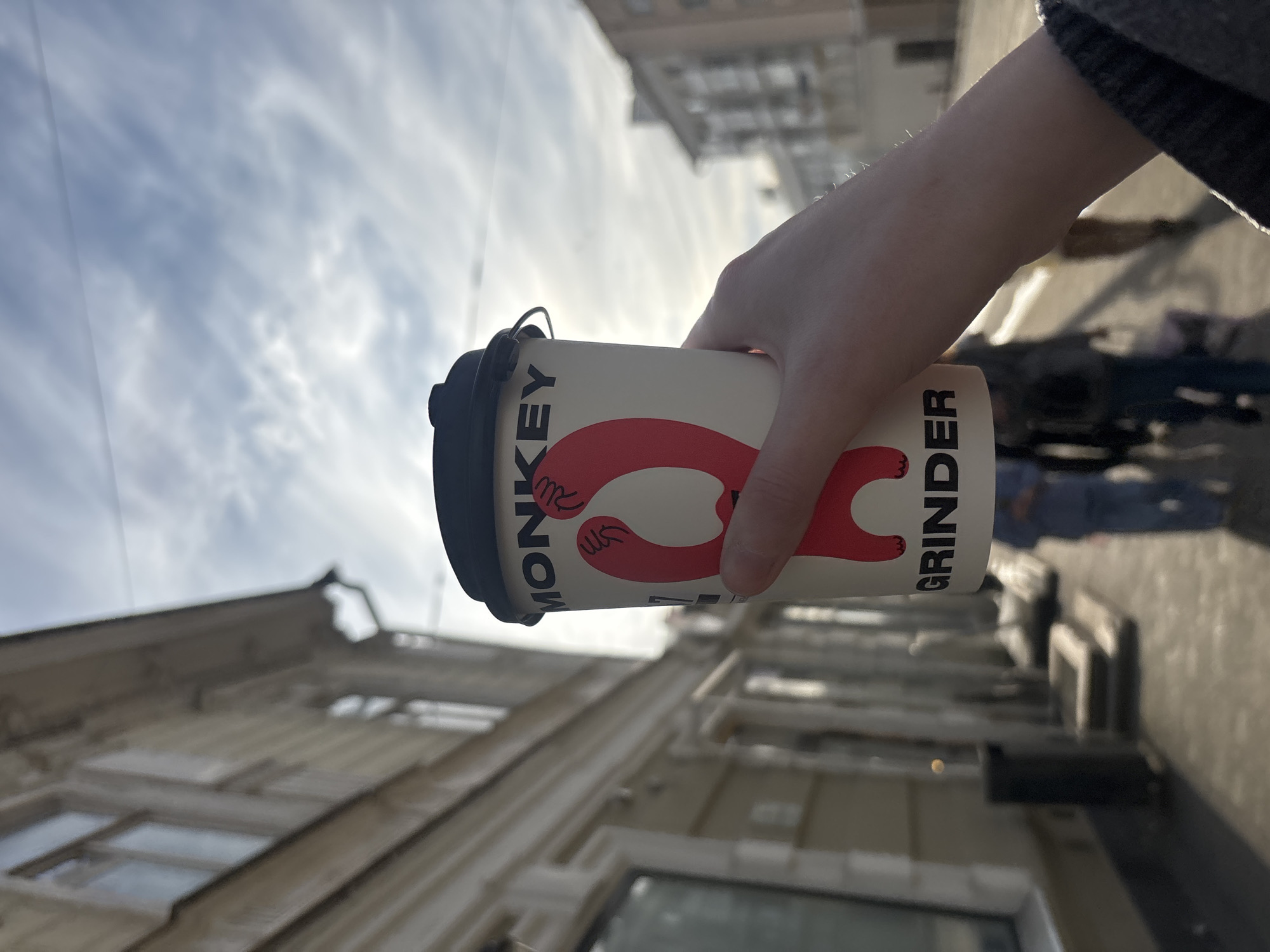 A person holds a coffee cup with "MONKEY GRINDER" printed on it. The background shows a street scene in Perm, Russia. Buildings are visible, along with blurry figures of people walking on the sidewalk. The sky is overcast with a thin layer of clouds.