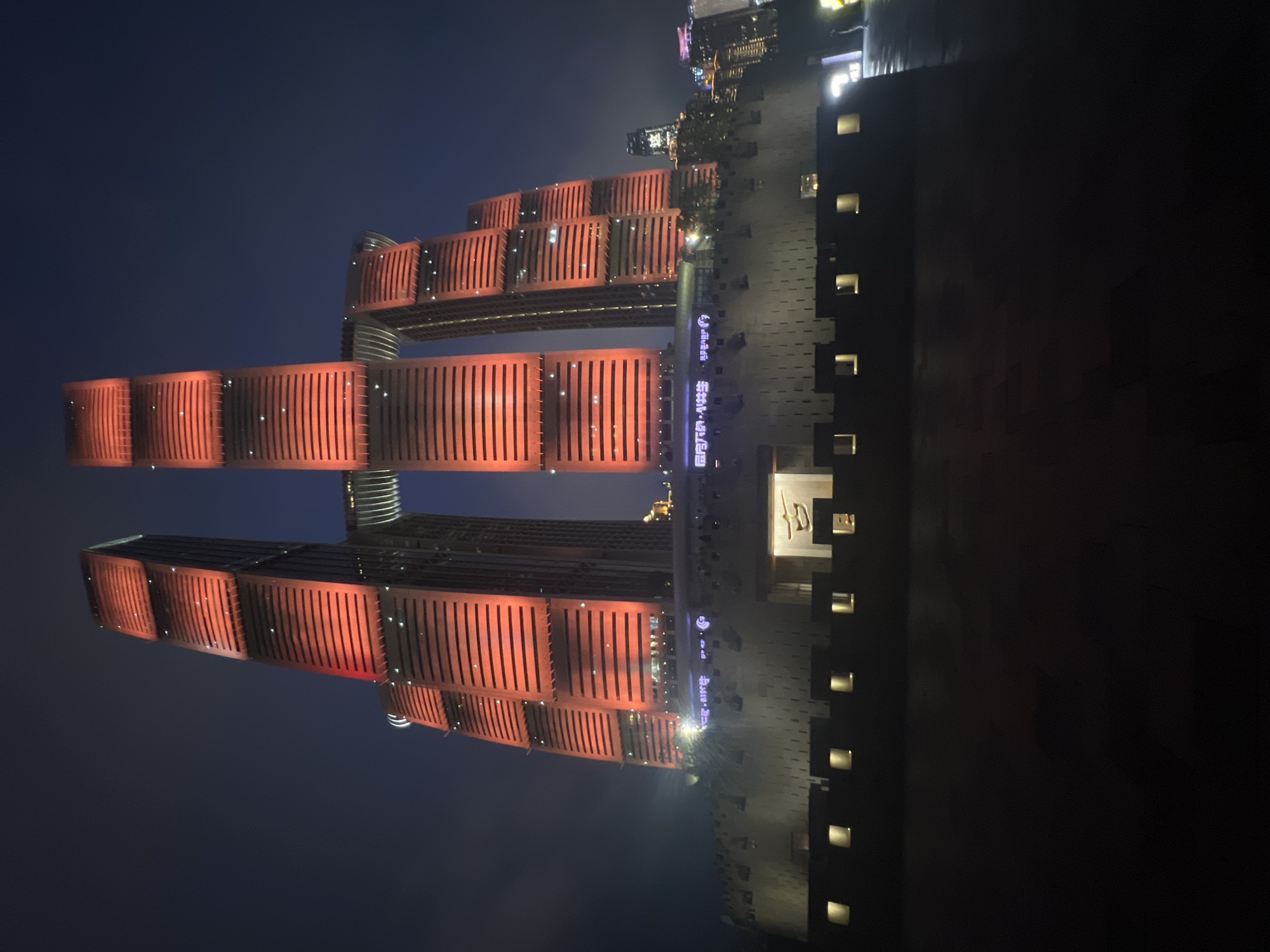 The image shows a night view of the Raffles City Chongqing complex in Yuzhong District, China. The complex is characterized by its distinctive architecture, featuring two towering skyscrapers connected by a horizontal skybridge. The buildings are illuminated with warm red and orange lights, highlighting their segmented facades. In the foreground, a textured, fortress-like base surrounds the skyscrapers, with small, illuminated rectangular windows. The sky is dark, and the overall scene conveys a sense of urban grandeur and nighttime illumination.